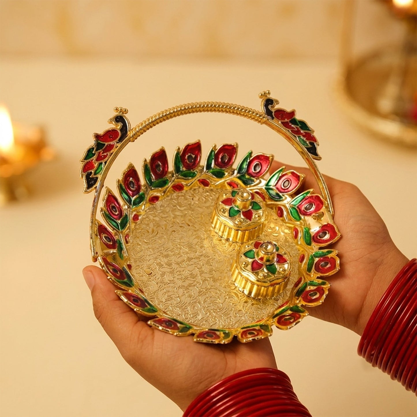 Hands holding a golden kumkum dish with a peacock handle and two lidded containers, with a blurred background.