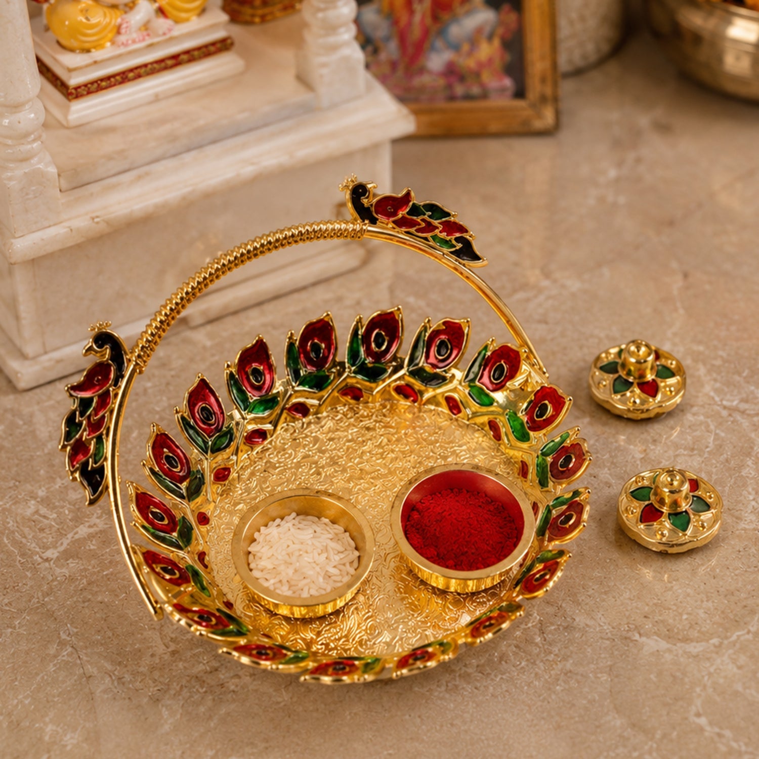 A golden kumkum dish with a peacock handle, holding red kumkum and white rice, with two lidded containers, on a textured