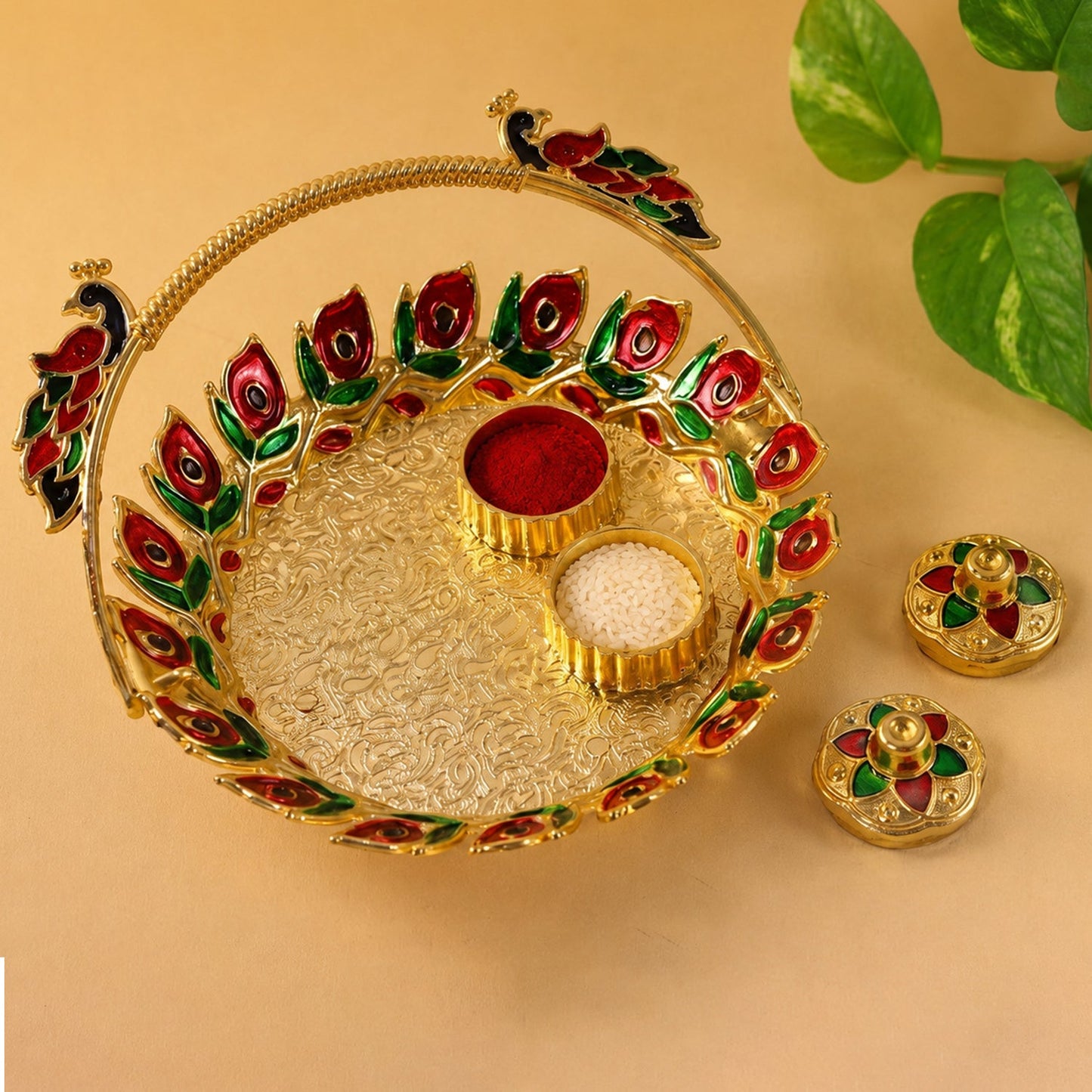 A golden kumkum dish with a peacock handle, holding red kumkum and white rice, with two lidded containers on a light brown