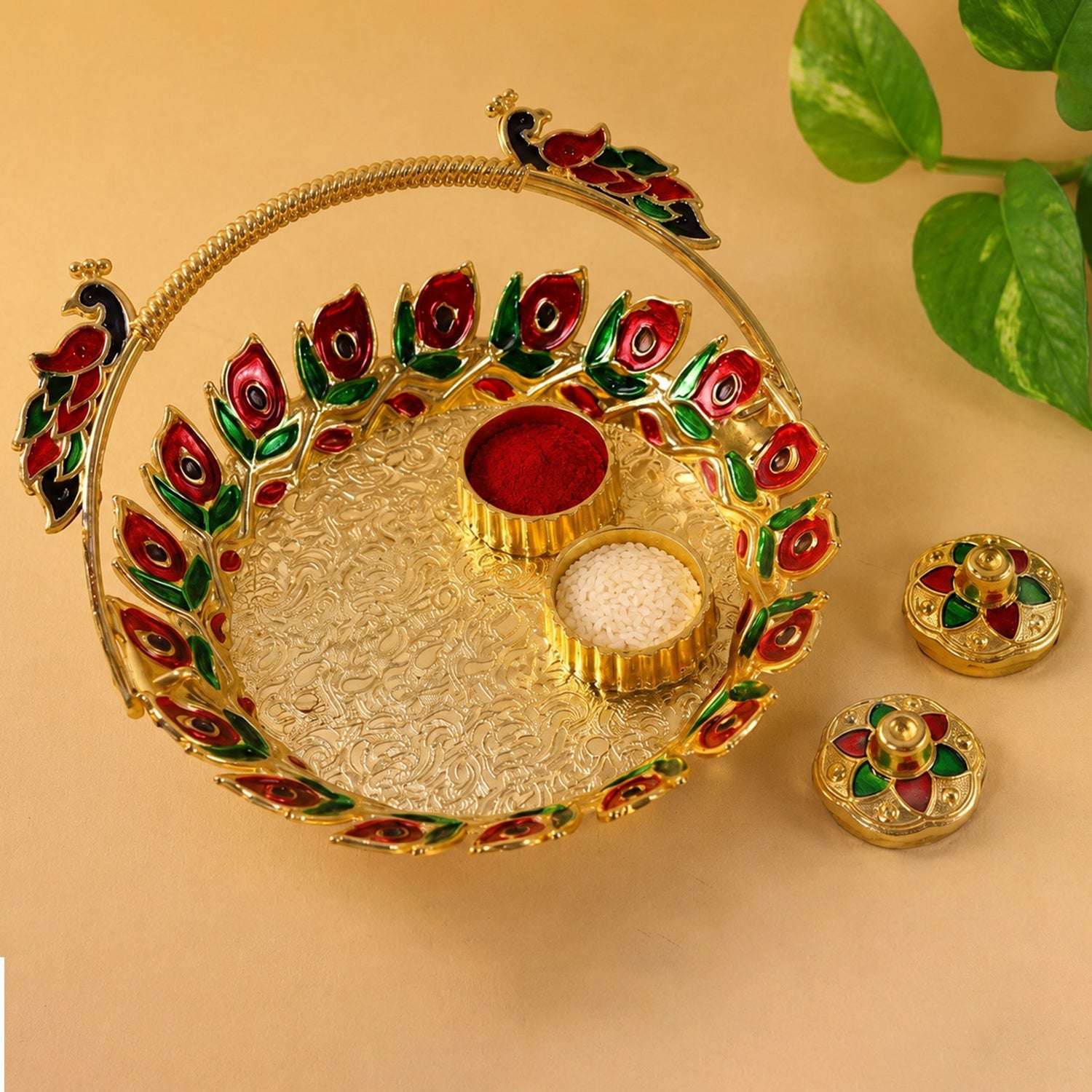 A golden kumkum dish with a peacock handle, holding red kumkum and white rice, with two lidded containers on a light brown