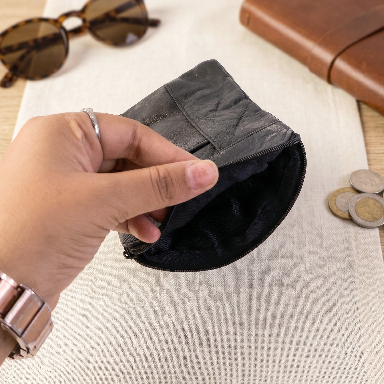 A hand opening a black leather pouch revealing its dark fabric lining, with sunglasses and coins nearby.