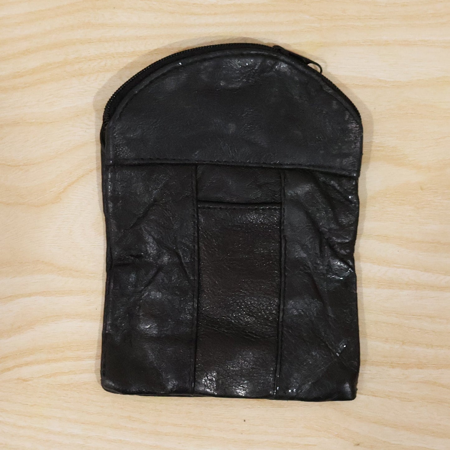 A black leather pouch with a top zipper and vertical seam details, laid flat on a light wooden surface.