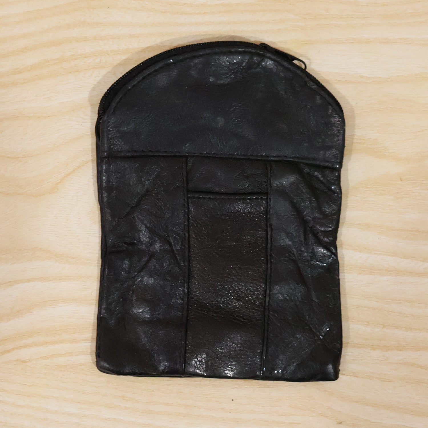A black leather pouch with a top zipper and vertical seam details, laid flat on a light wooden surface.