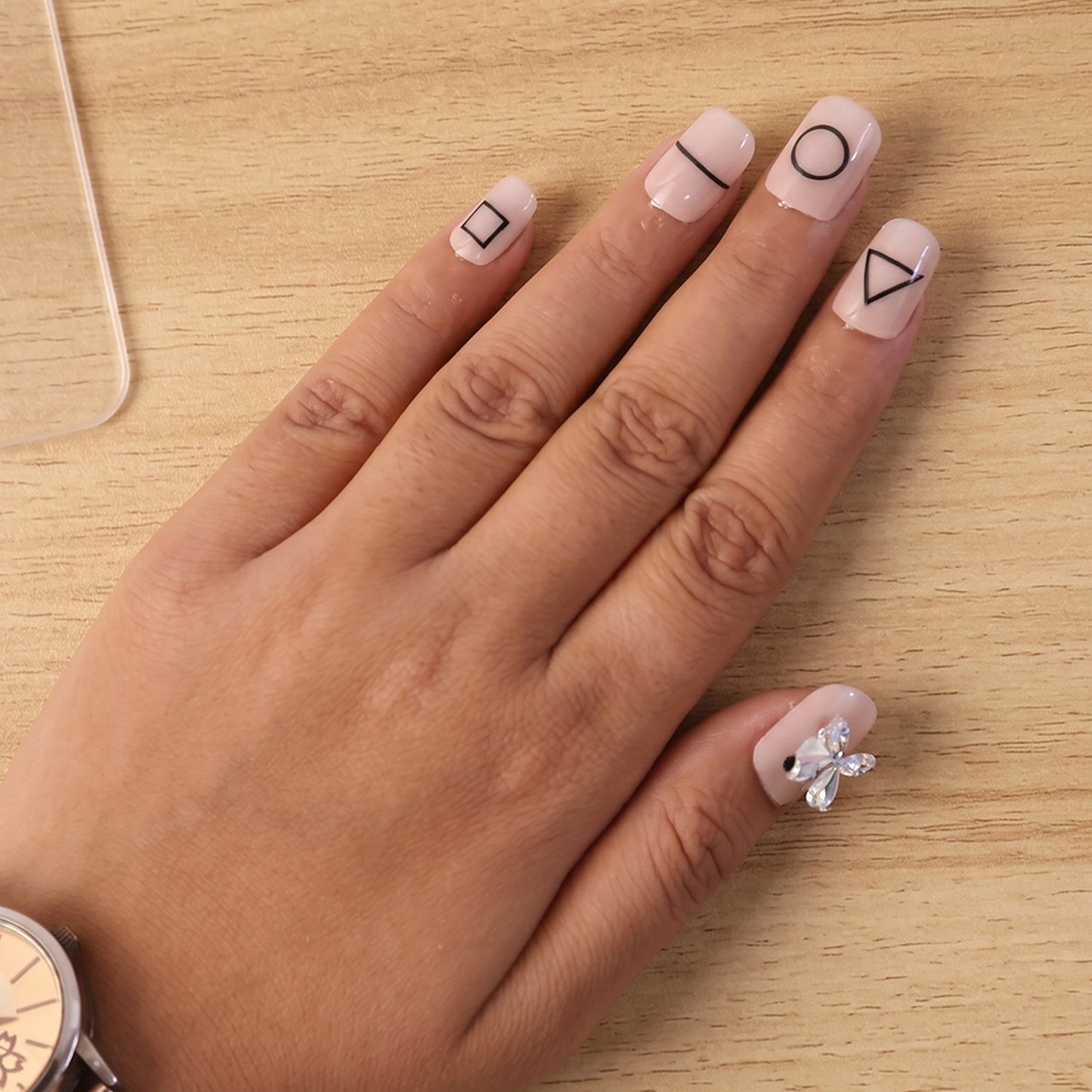 A person's hand wearing nude press-on nails with black geometric shapes and one iridescent bow, next to a watch.