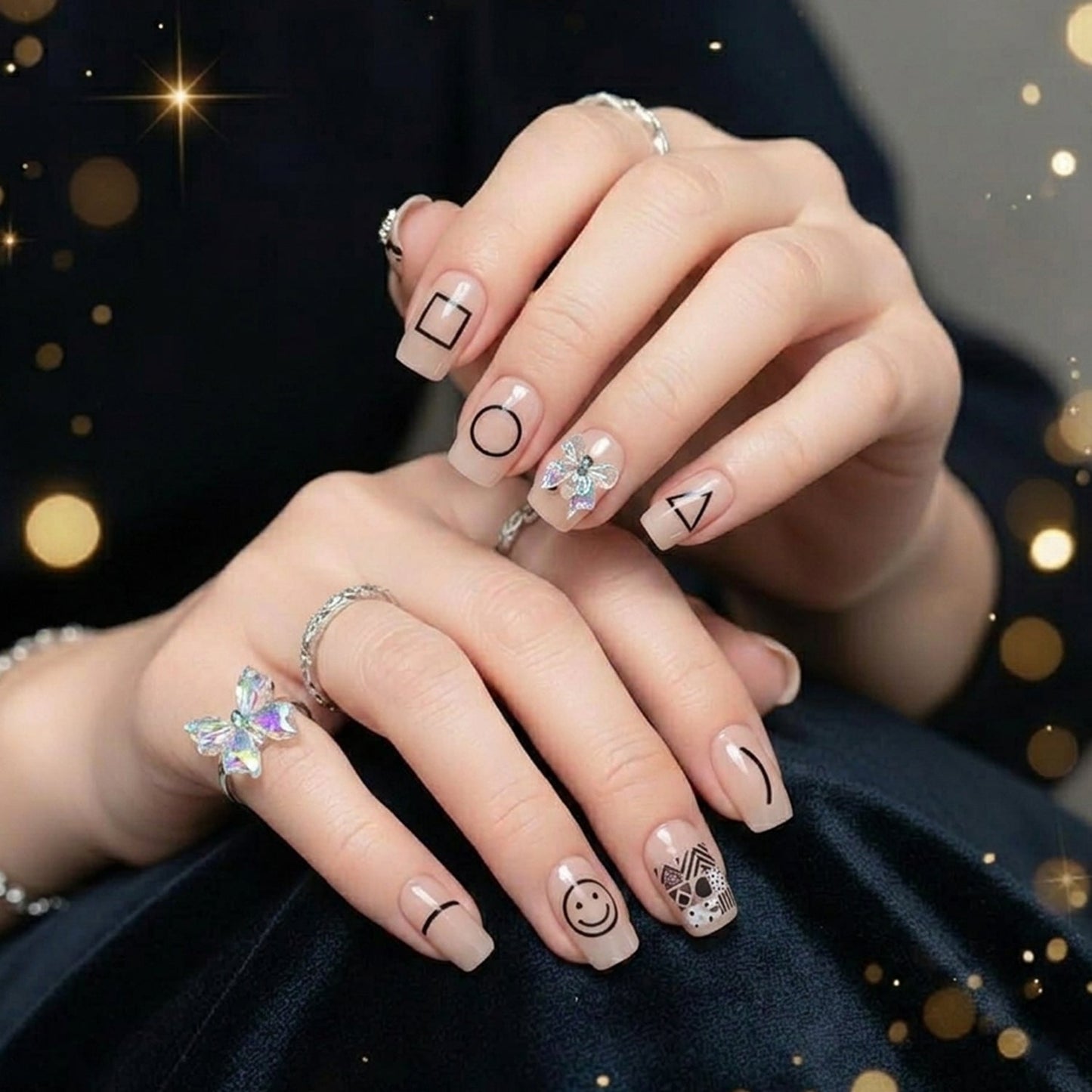 A person's hands wearing nude press-on nails with black geometric shapes, one with an iridescent bow, against a dark