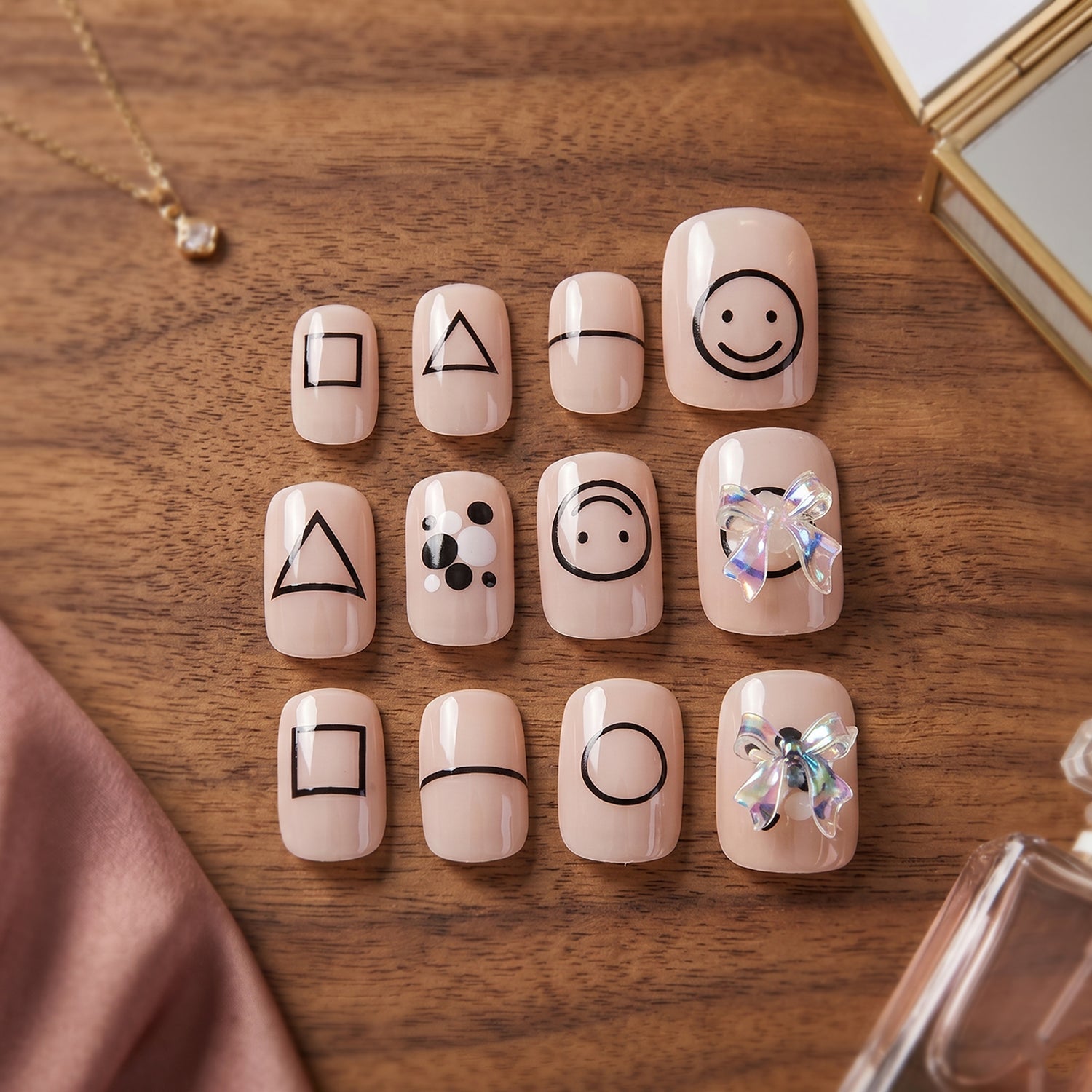 A set of 12 nude press-on nails with black geometric shapes and two iridescent bows, on a wooden surface with accessories.
