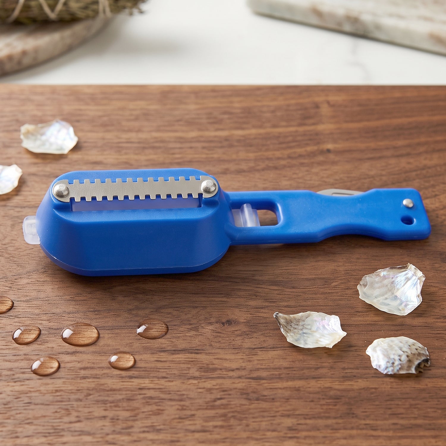 The blue fish scaler with a metal blade is packaged in a red and white blister pack, displayed on wood.
