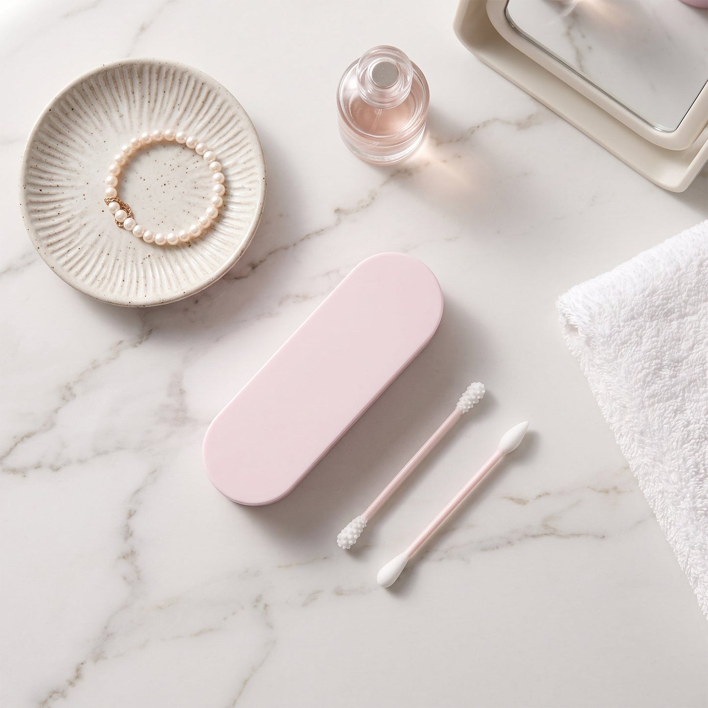 Pink reusable silicone ear swab case and two swabs on a marble surface with a pearl bracelet, perfume, and white towel.