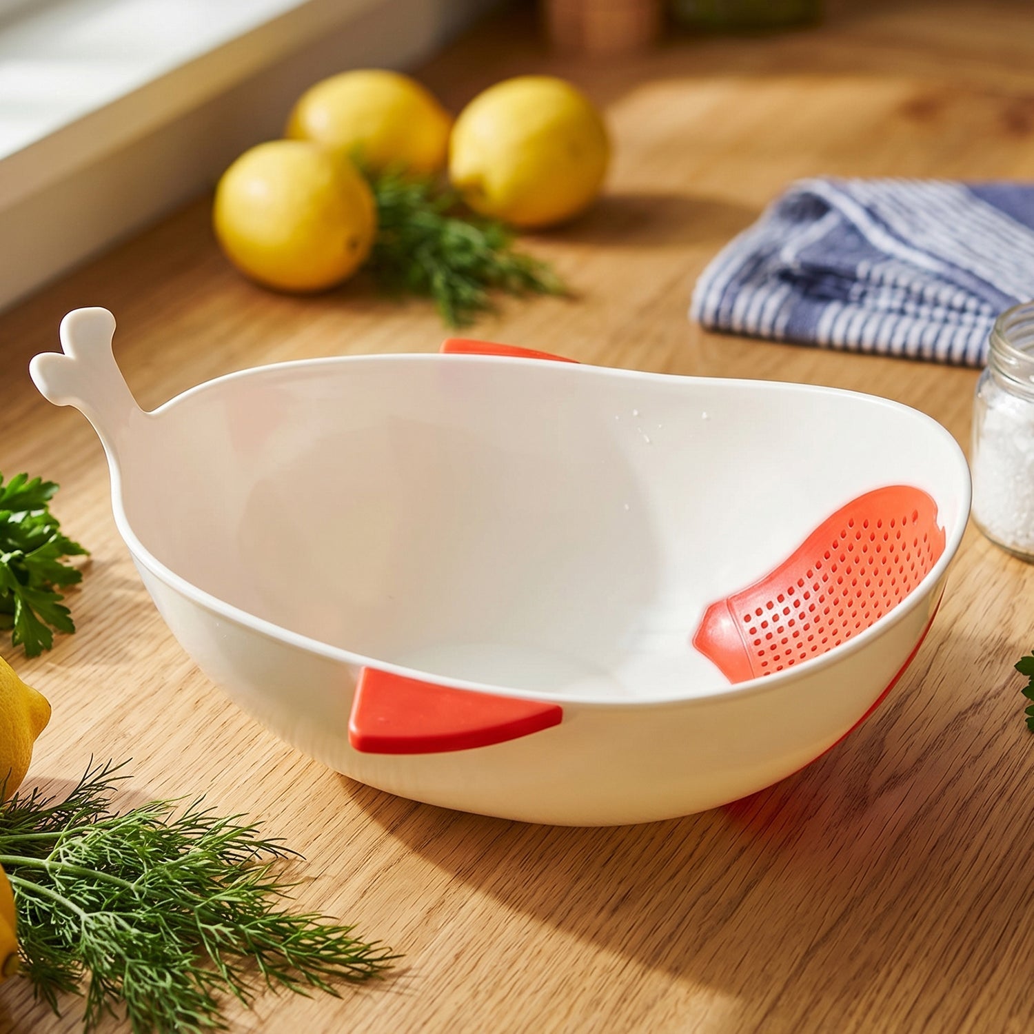 A white whale-shaped rice drainer bowl with red accents sits on a wooden counter next to lemons and fresh dill.