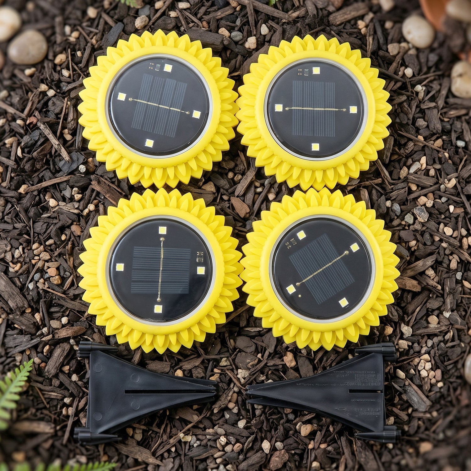 Four yellow, sunflower-shaped solar lights with black solar panels and two black ground stakes are arranged on dark soil.