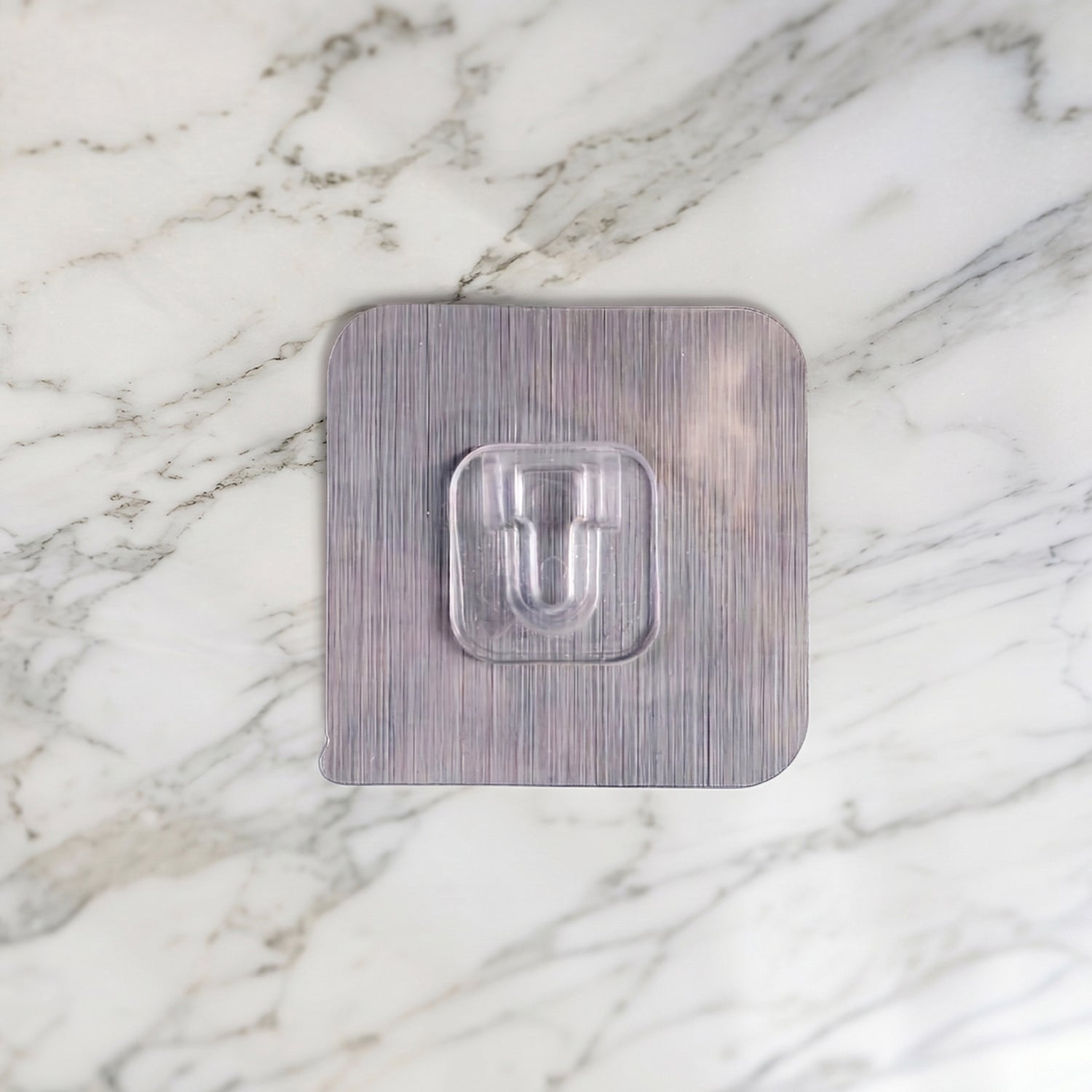 A transparent self-adhesive hook with a brushed metal-look backing centered on a white marble background.