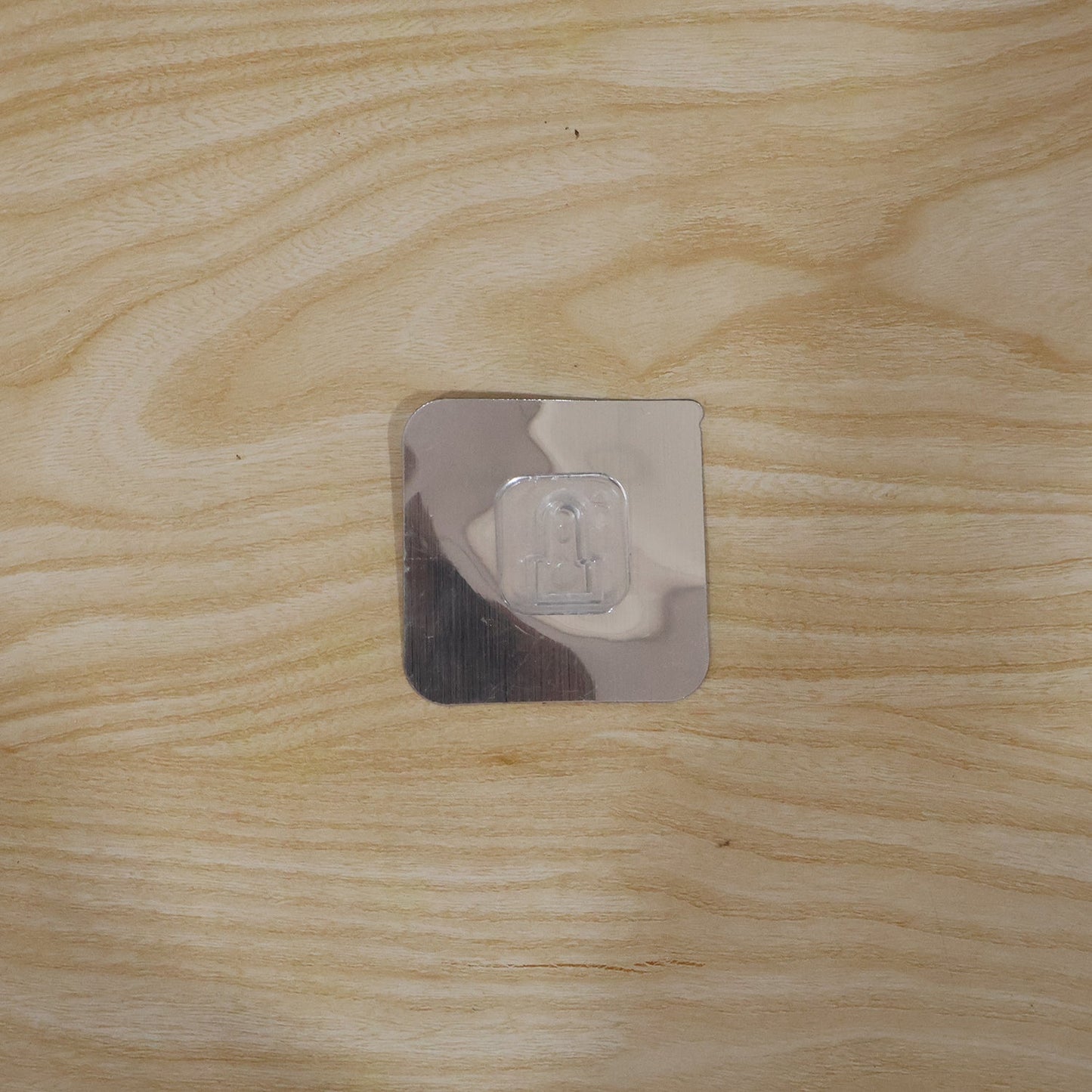 A single transparent self-adhesive hook with a reflective backing adhered to a light wooden surface.