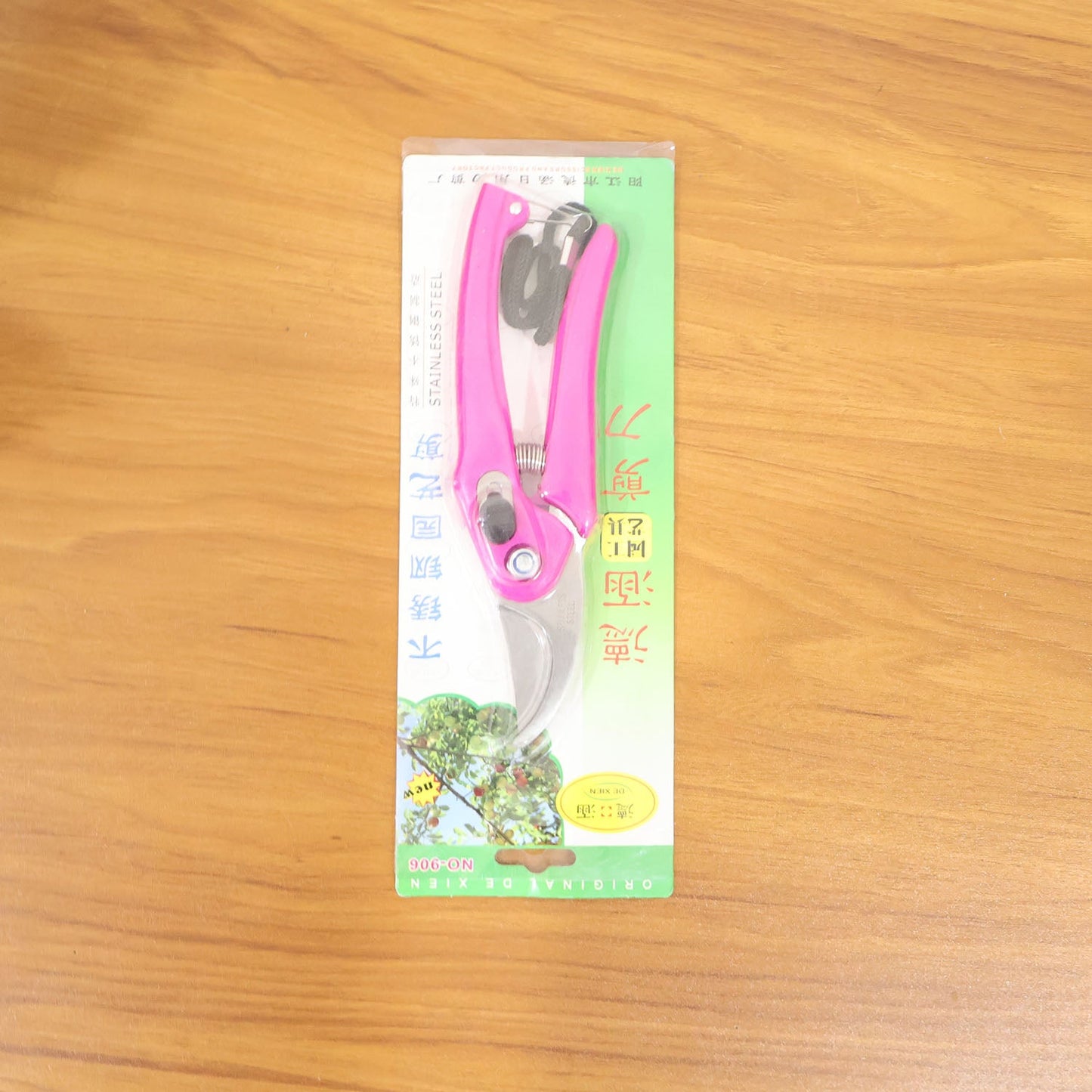 Pink pruning shears packaged in clear plastic with green and yellow accents, lying on a light brown wooden surface.