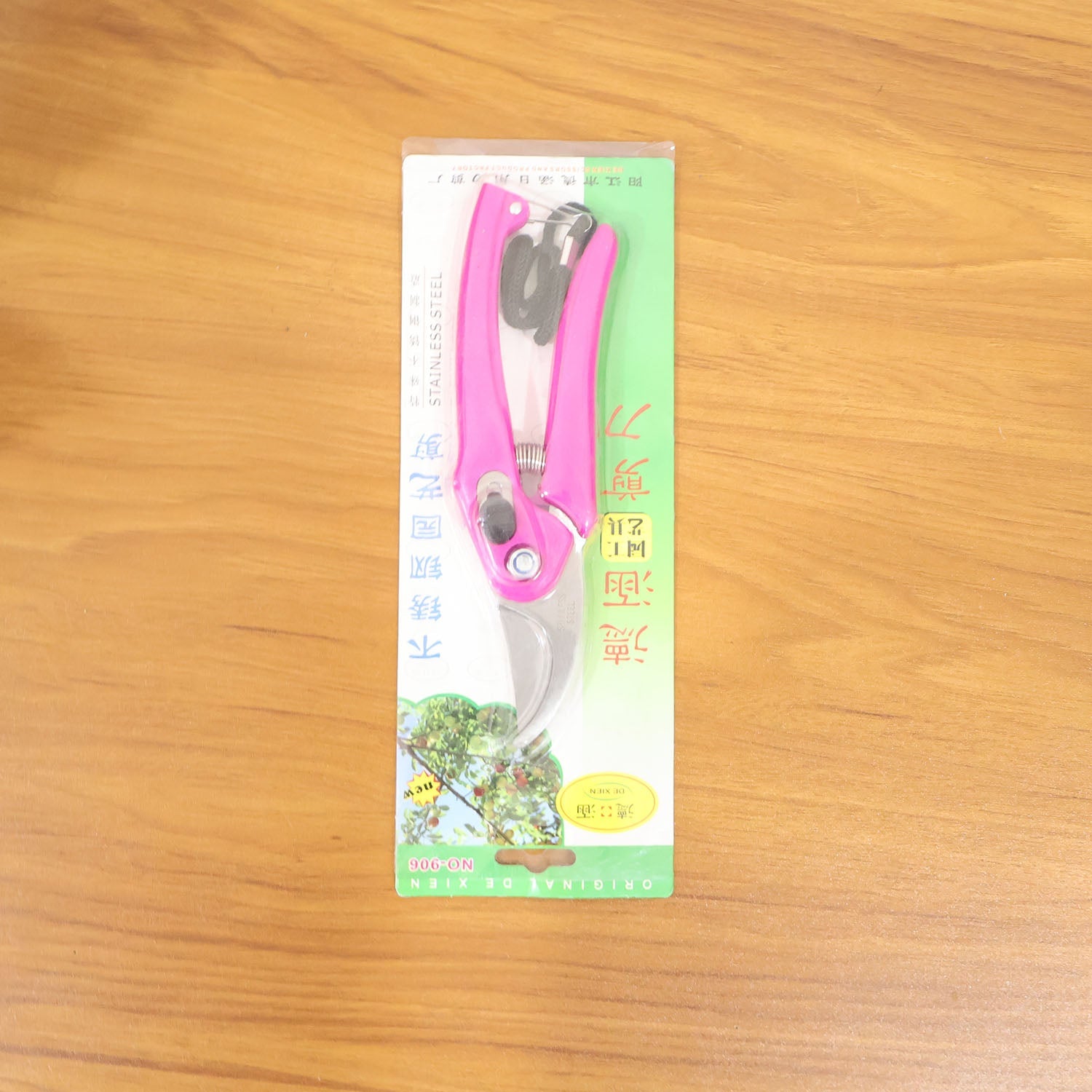 Pink pruning shears packaged in clear plastic with green and yellow accents, lying on a light brown wooden surface.