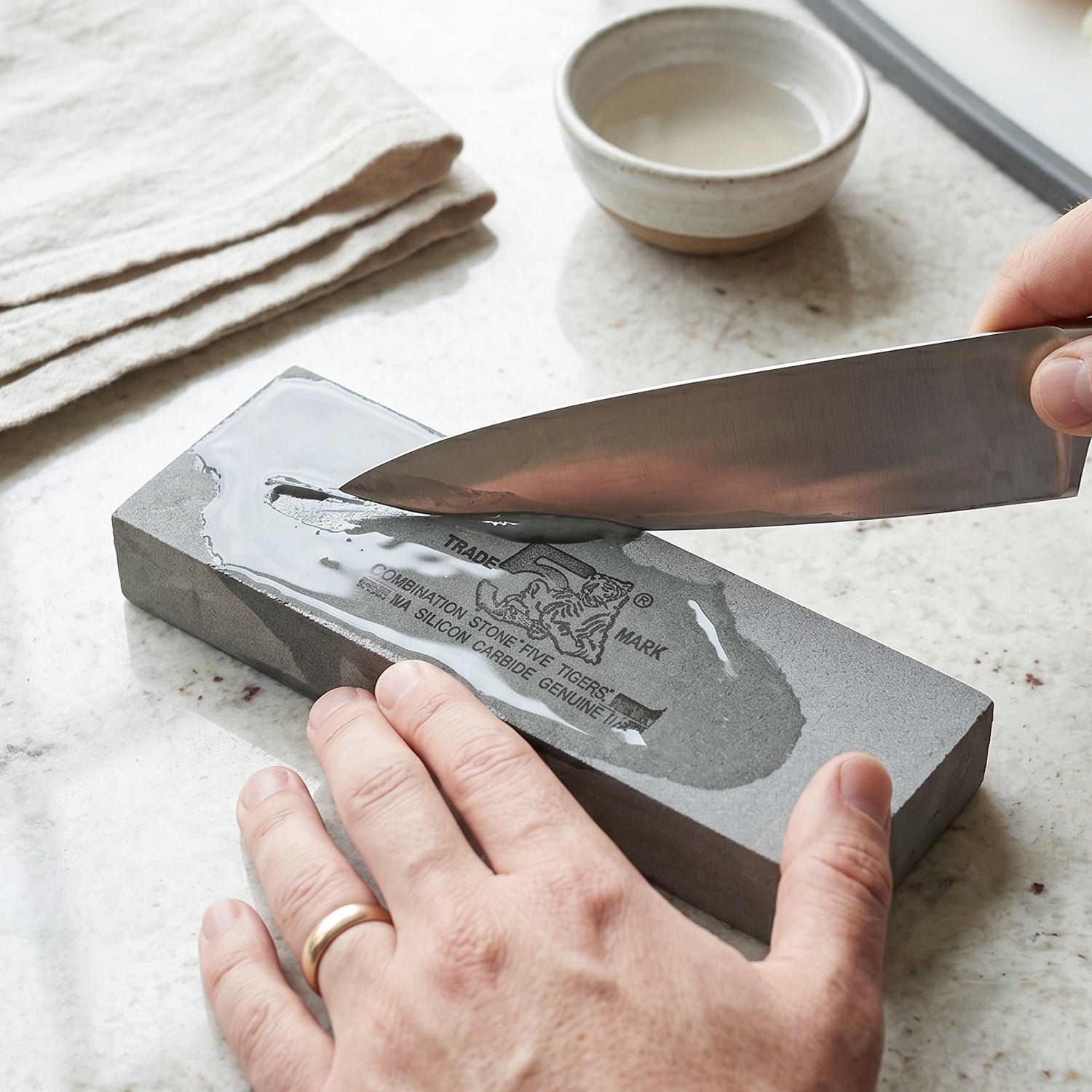 Hands sharpening a silver knife on a wet gray stone with a tiger logo, next to a small bowl of water.