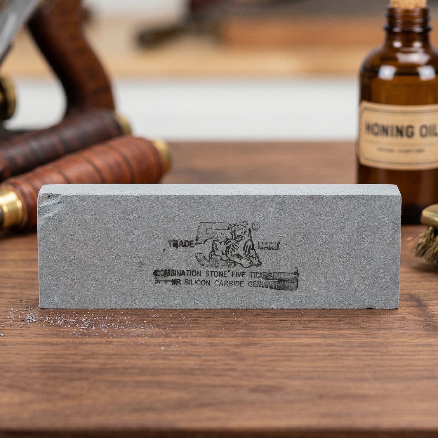 Gray rectangular sharpening stone with black text and tiger logo, on a wooden surface with honing oil and tools in