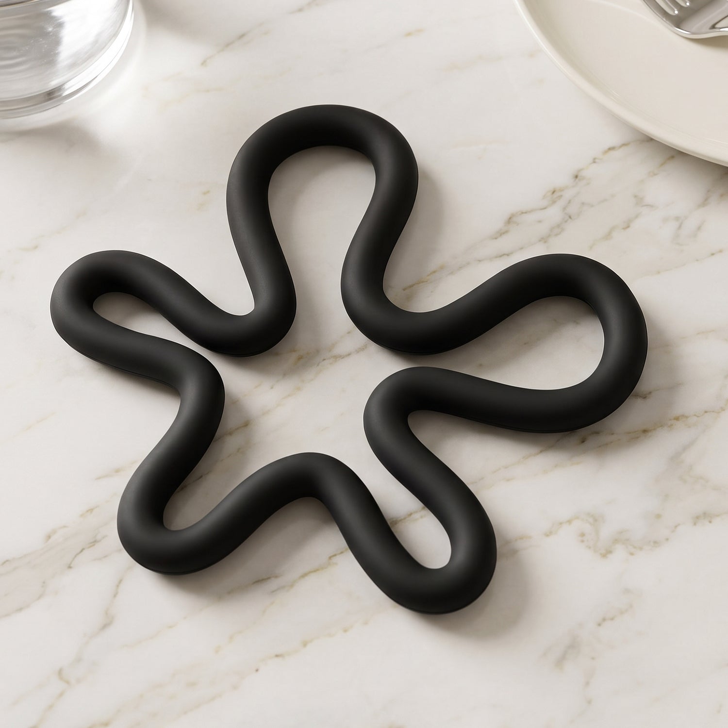 A black silicone flower-shaped hot pot holder on a white marble surface next to a clear glass and a white plate.