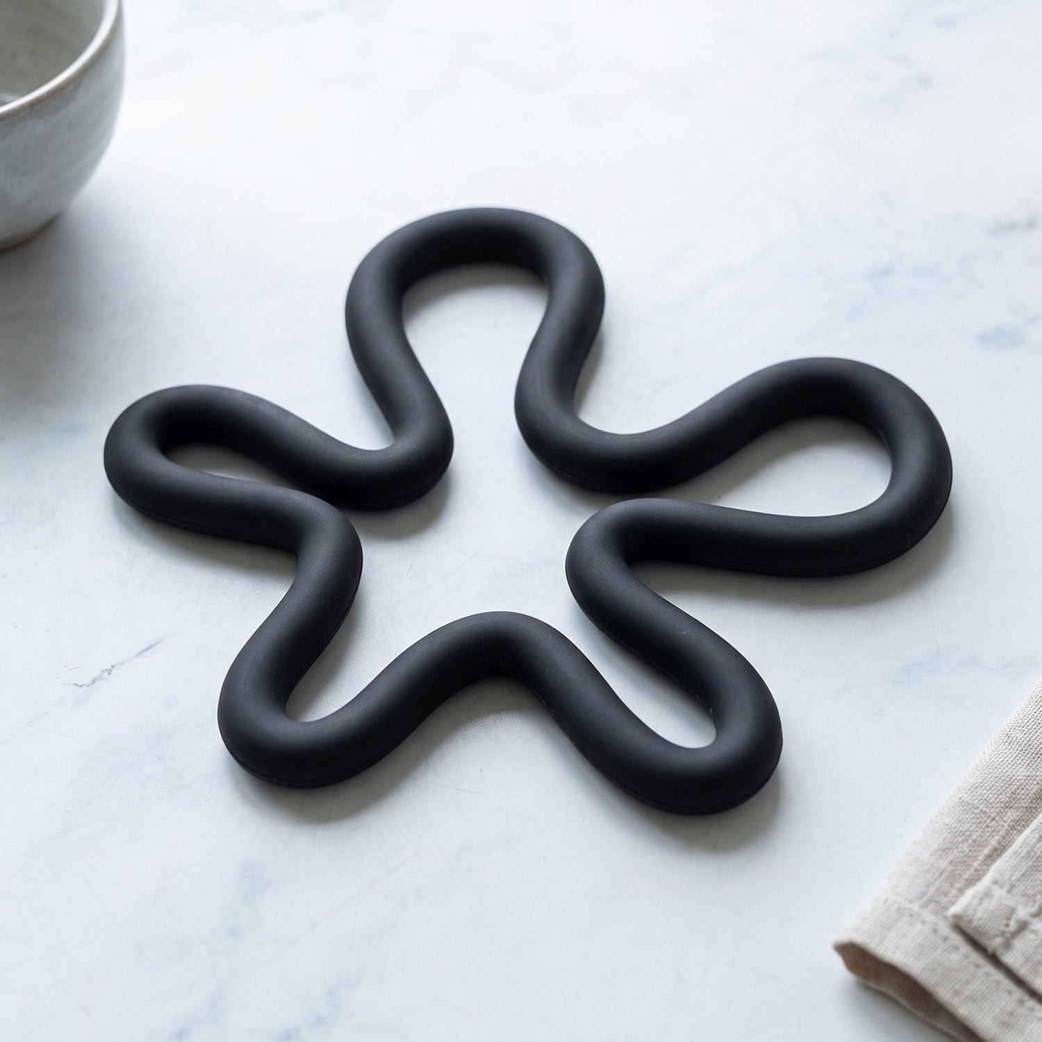 A black silicone flower-shaped hot pot holder on a white marble surface next to a gray bowl and a beige linen napkin.