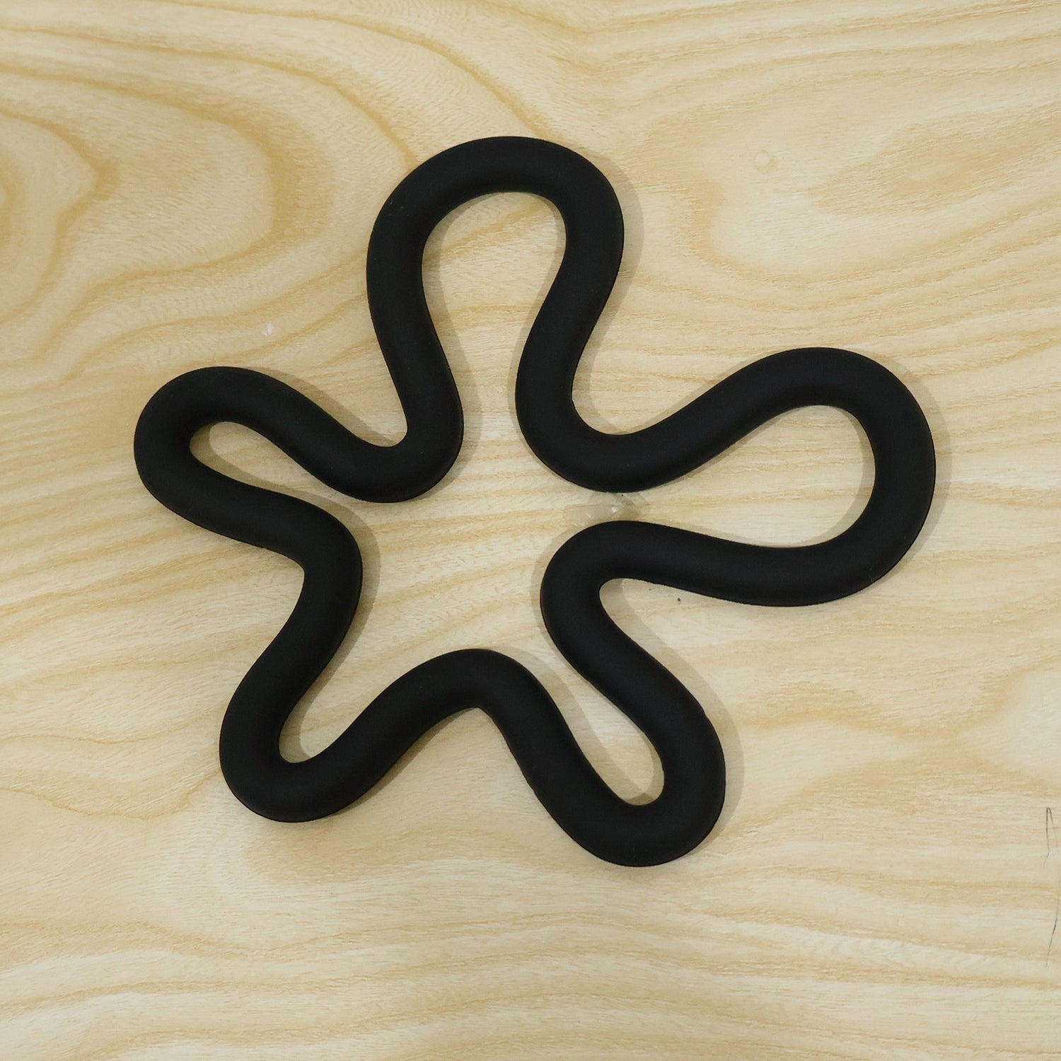 A black silicone flower-shaped hot pot holder on a light wooden surface, viewed from directly above.