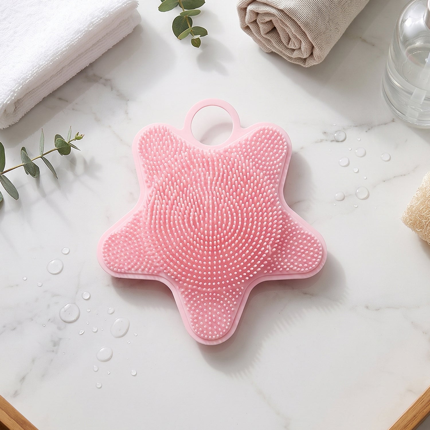 A pink silicone starfish-shaped brush rests on a white marble surface with towels, a plant, and a soap dispenser in the