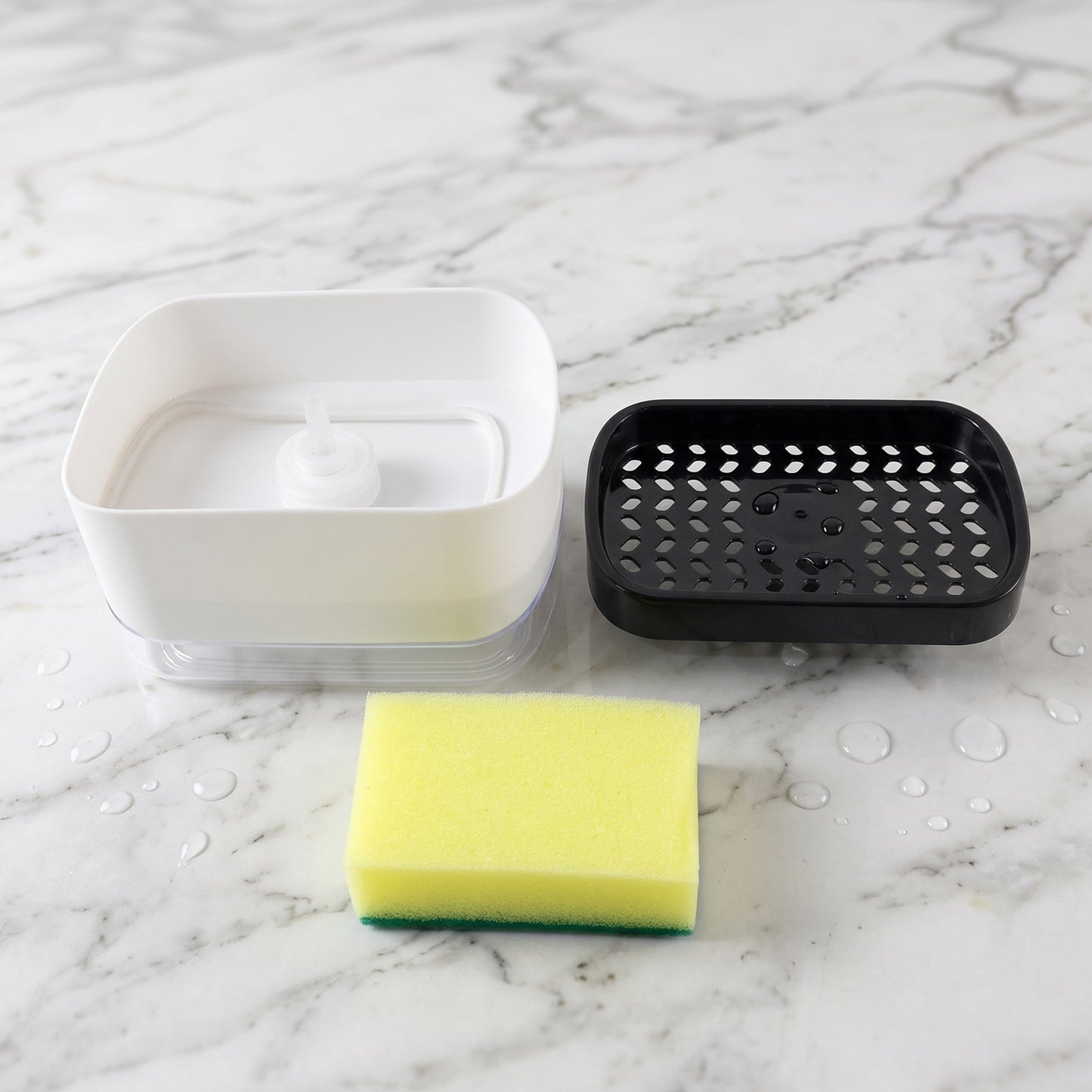 The white soap dispenser base, black perforated top, and yellow sponge are separated on a white marble surface with water