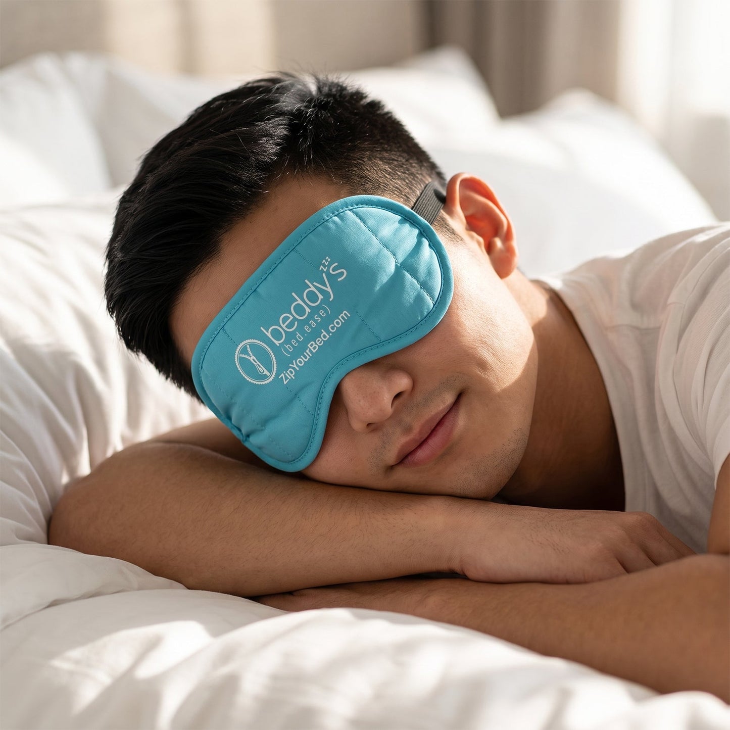 A man with short dark hair sleeps on a white pillow, wearing a light blue quilted sleep mask with a white logo.