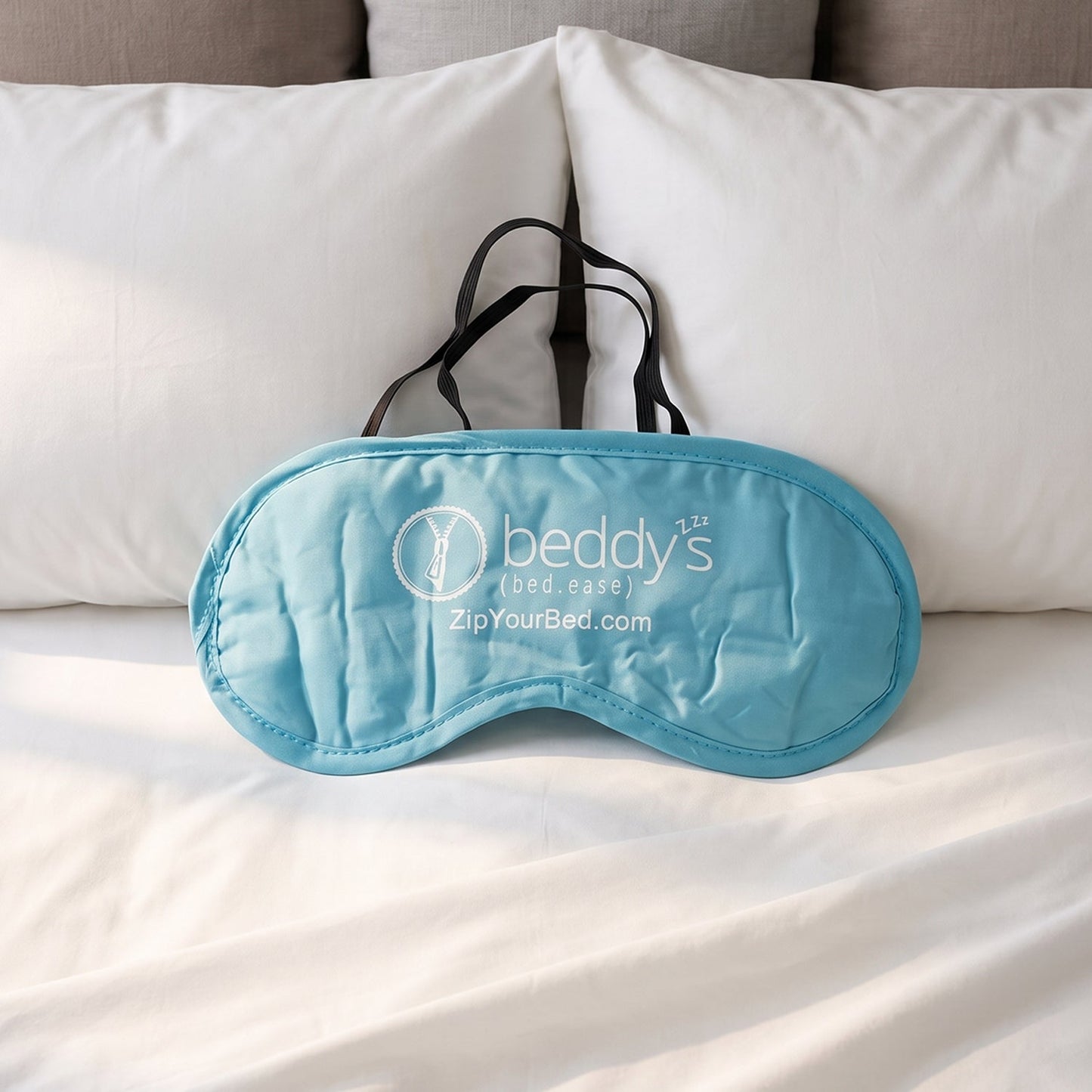 A light blue sleep mask with a white logo and black elastic strap rests on a neatly made white bed with pillows.