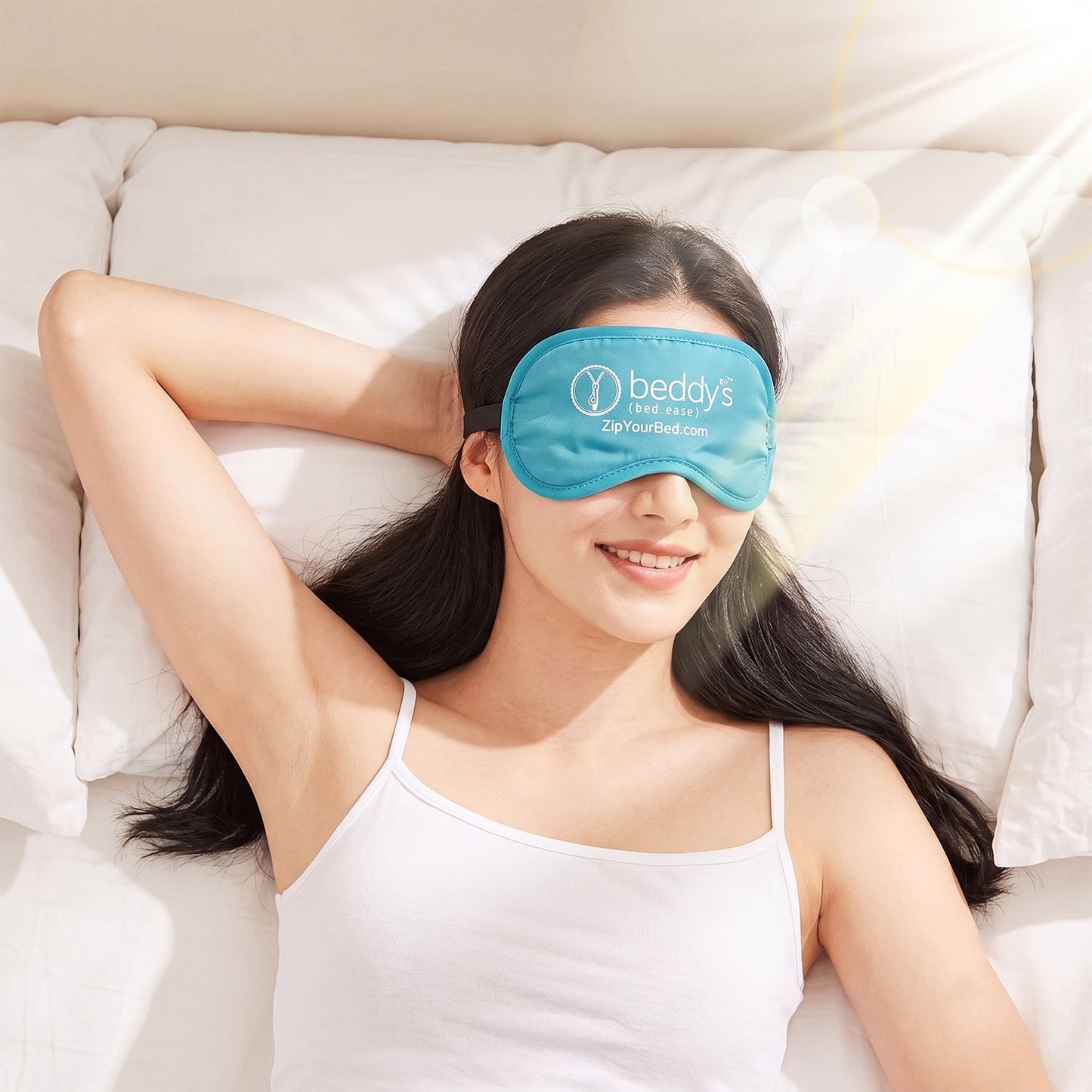 A woman in a white tank top smiles while lying on a white bed, wearing a light blue sleep mask with a white logo.