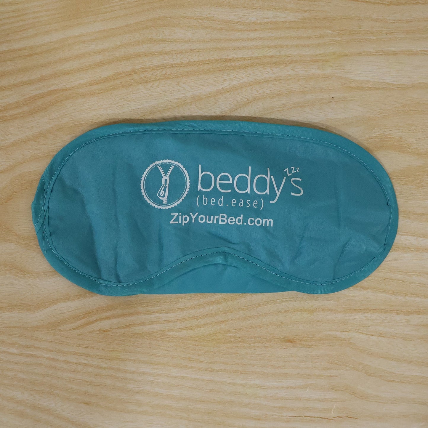 A light blue sleep mask with a white logo is laid flat on a light brown wooden surface.