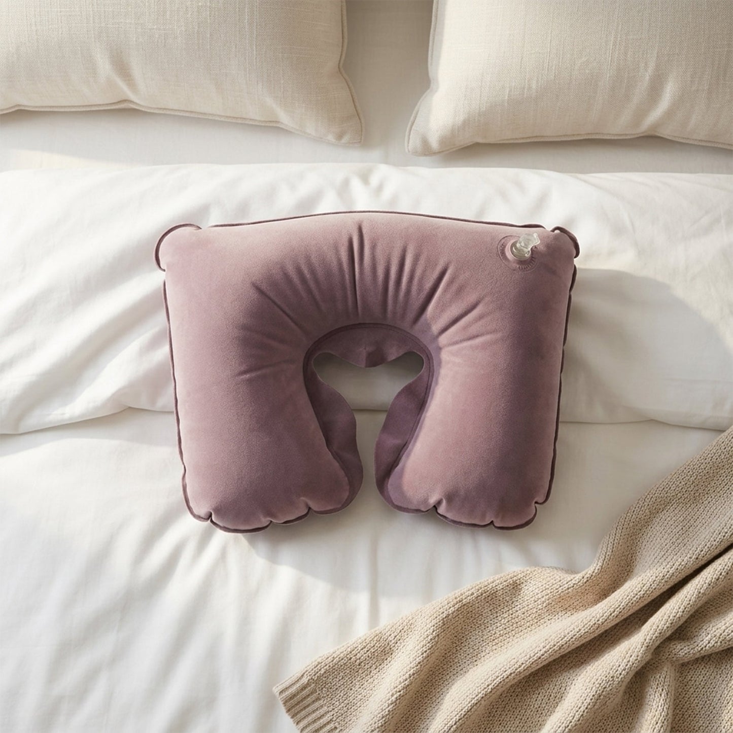 An inflated purple velvet U-shape neck pillow lies on a white bed with a beige blanket and pillows.