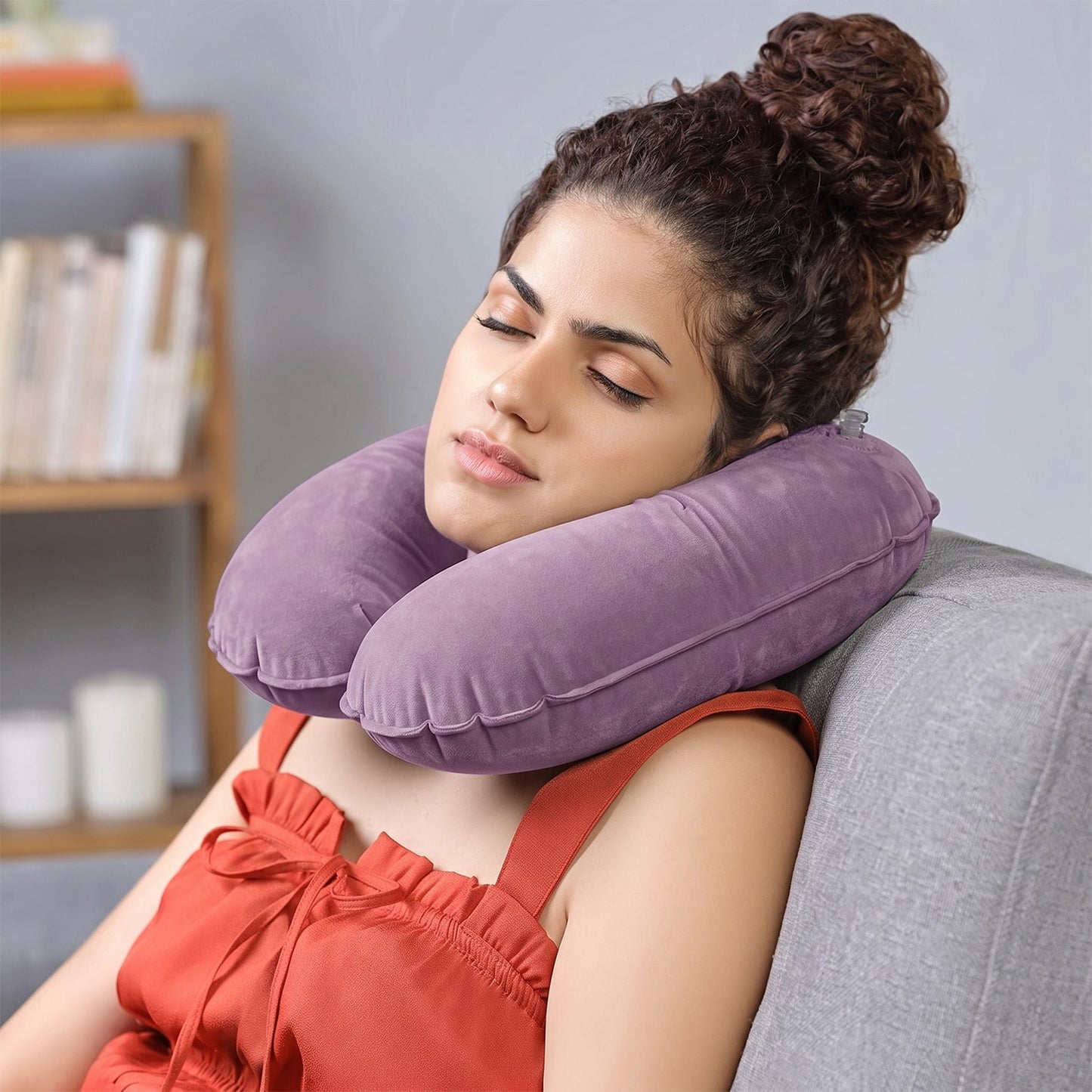 A woman with her eyes closed rests on a grey couch, wearing an inflated purple velvet U-shape neck pillow.