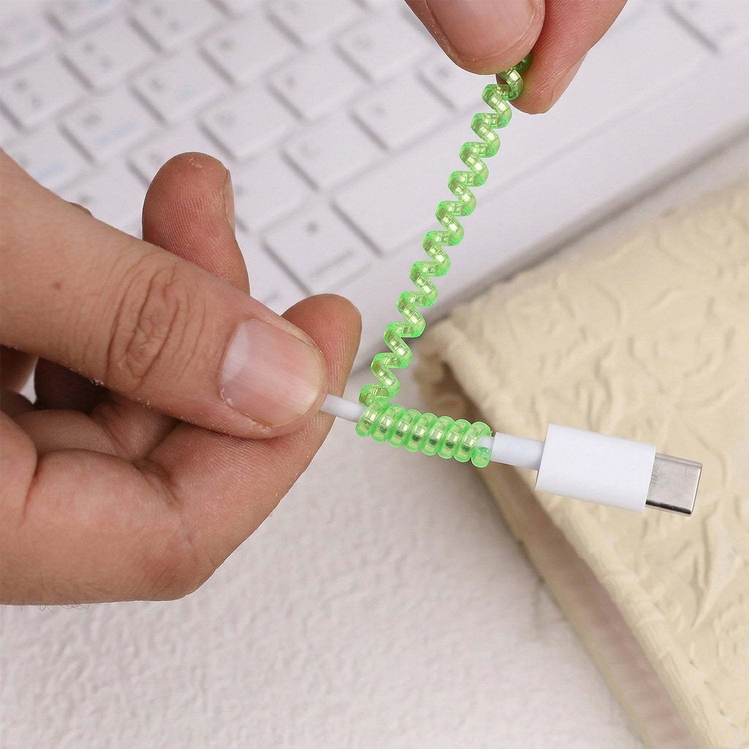Hands apply a translucent green spiral cable protector sleeve to a white charging cable over a keyboard.