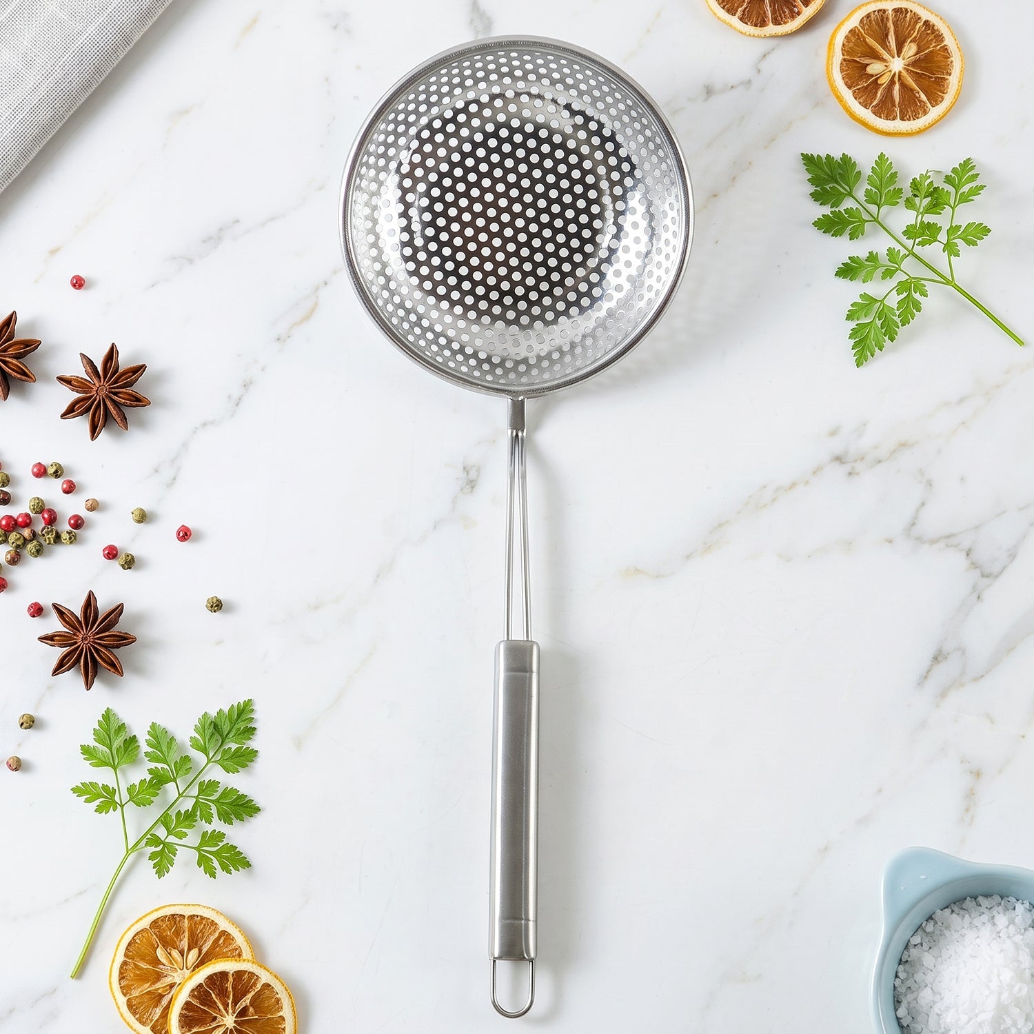 A stainless steel perforated skimmer spoon lies on a white marble surface surrounded by spices and herbs.
