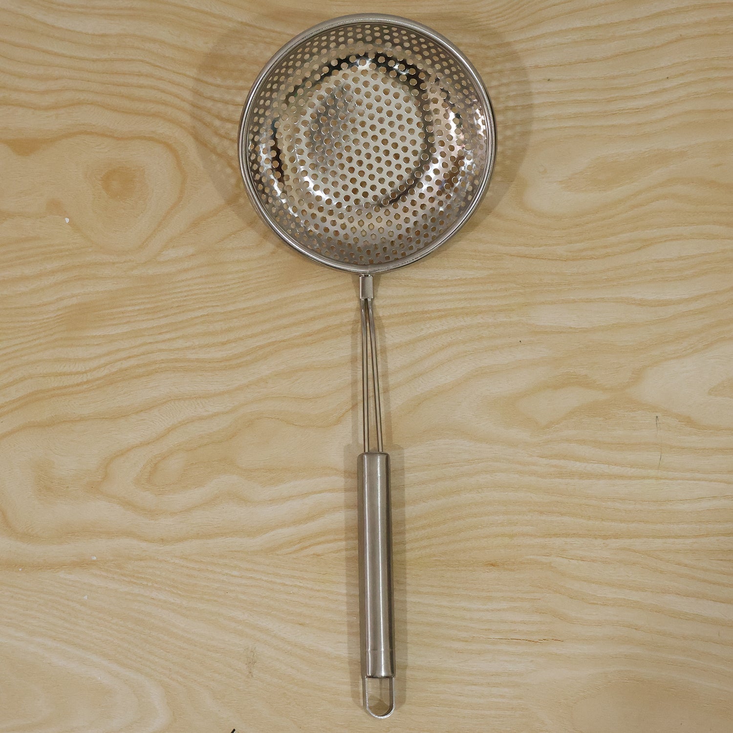 A stainless steel perforated skimmer spoon with a long handle resting on a light wooden surface.