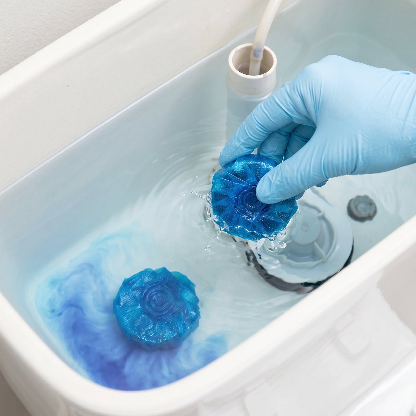 A gloved hand placing a blue toilet cleaner tablet into a toilet tank with blue water.