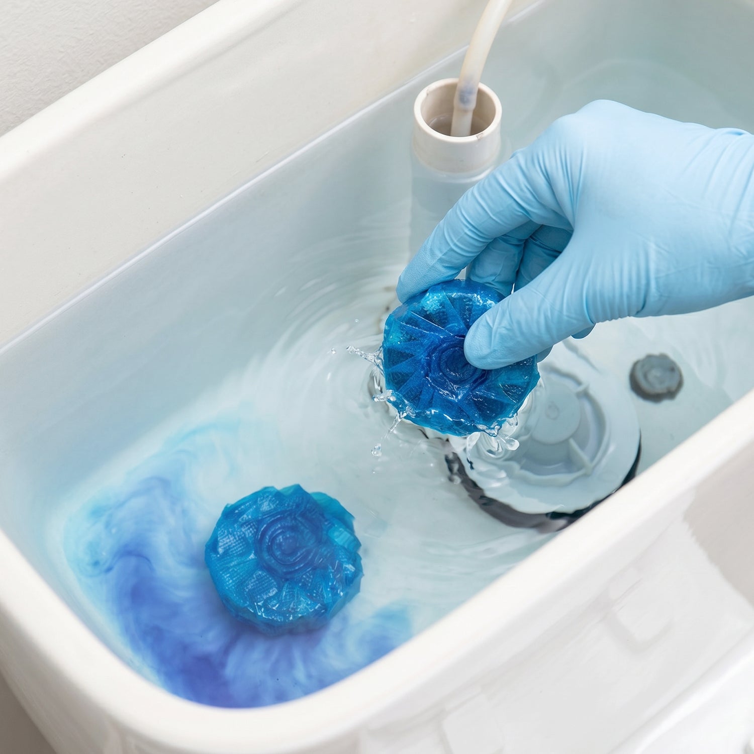 A gloved hand placing a blue toilet cleaner tablet into a toilet tank with blue water.