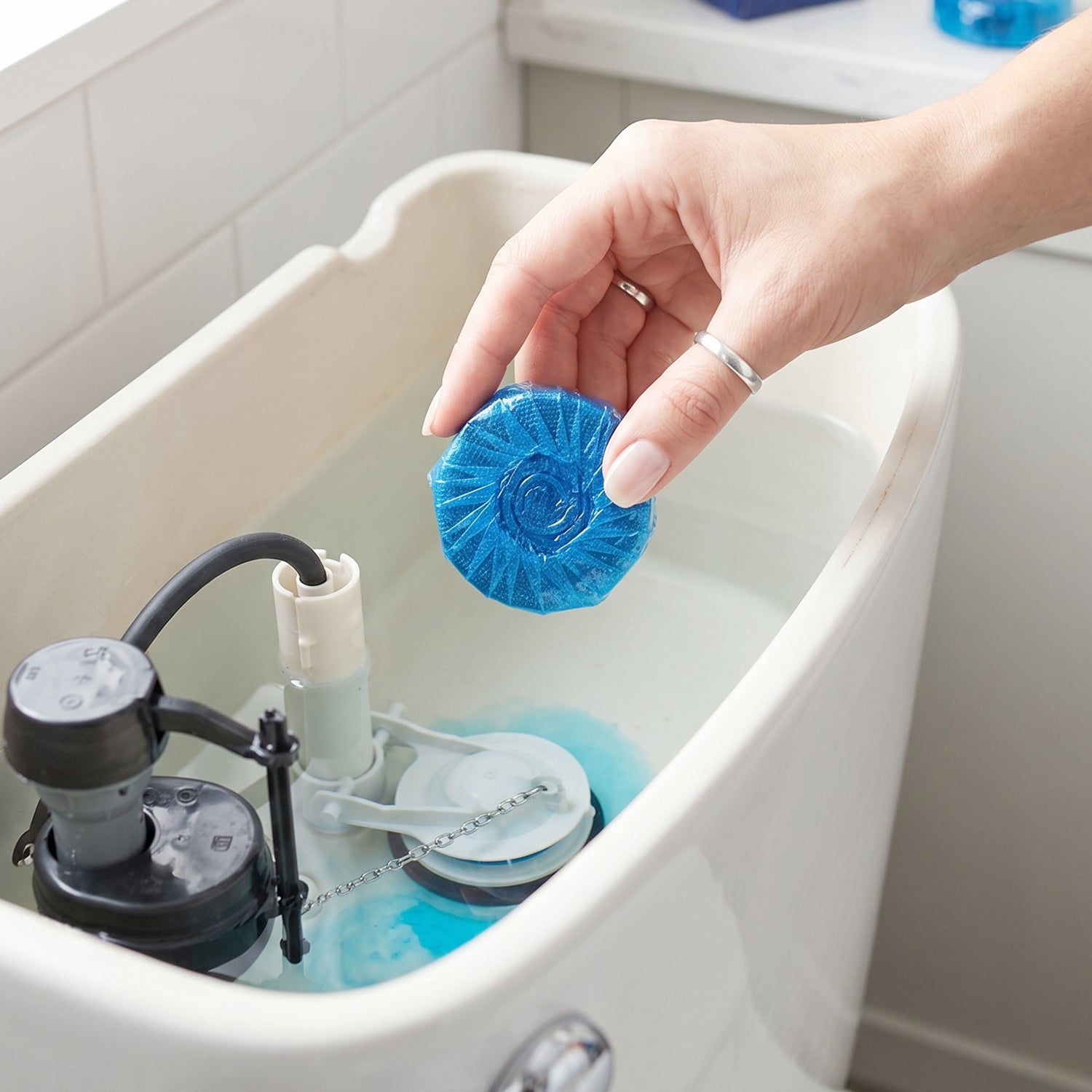 A hand placing a blue toilet cleaner tablet into a toilet tank with internal mechanisms visible.