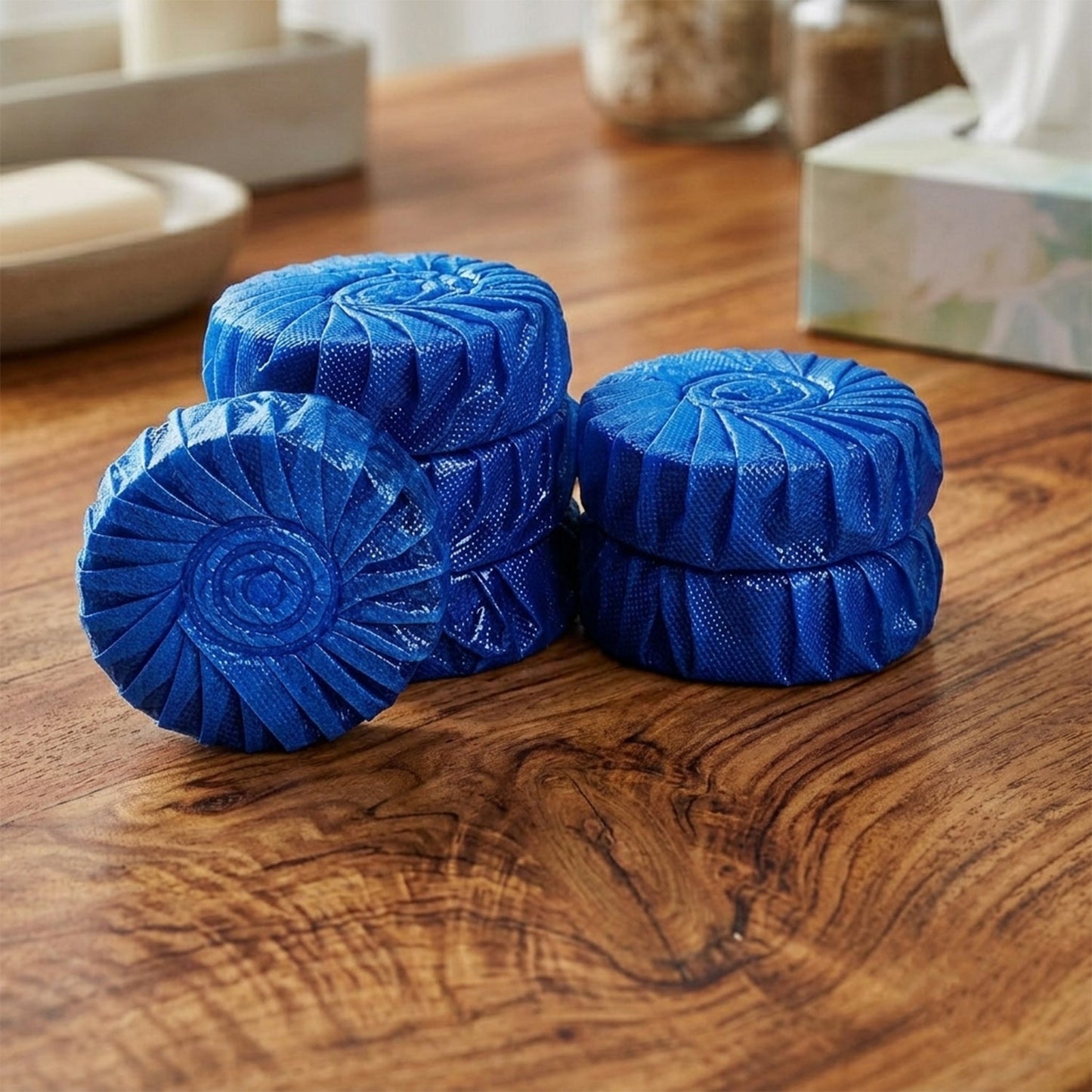 Six blue toilet cleaner tablets, some stacked, are arranged on a wooden surface with blurred bathroom items in the