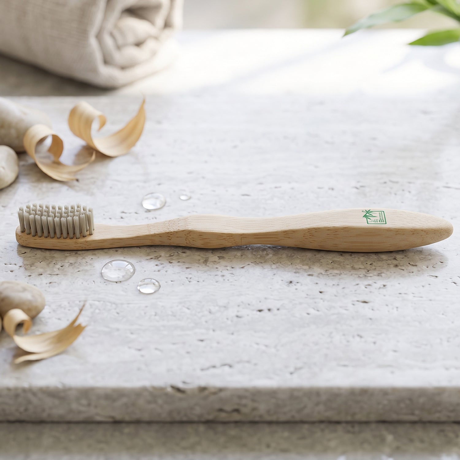 A single bamboo toothbrush with gray bristles lies on a light stone surface with water droplets and bamboo shavings.