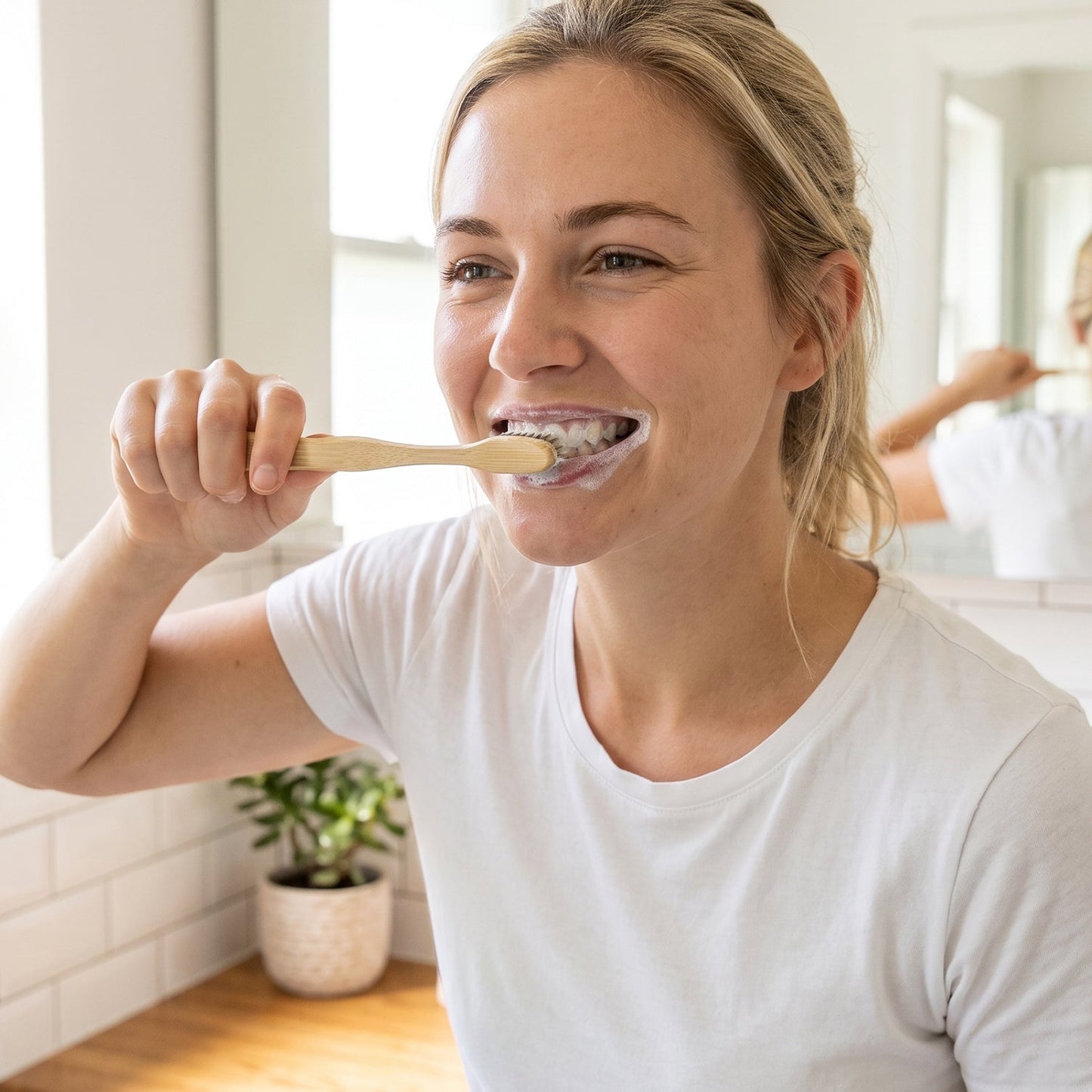 A smiling woman in a white t-shirt brushes her teeth with a bamboo toothbrush in a bathroom setting.