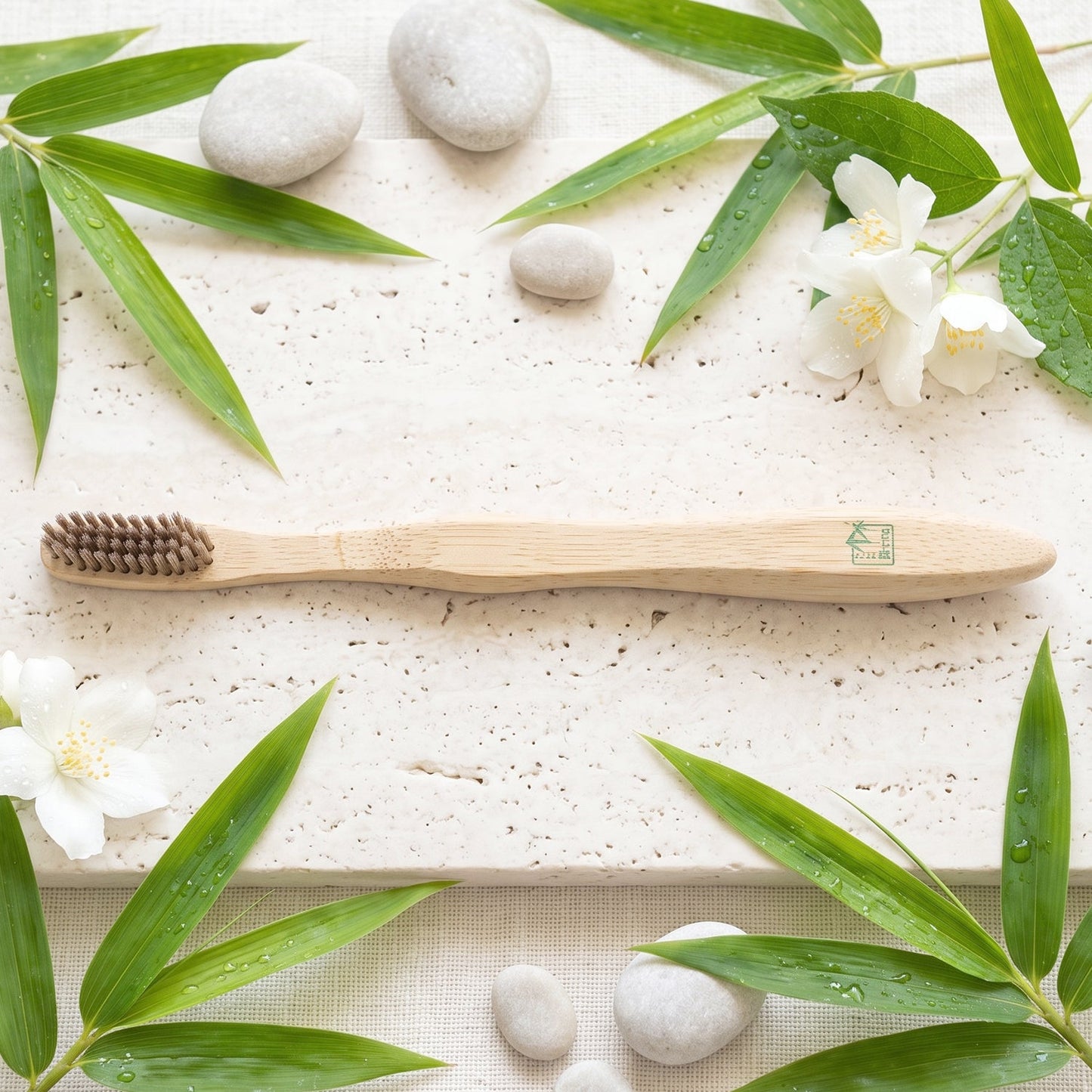 A bamboo toothbrush on a light stone surface, surrounded by green bamboo leaves, white flowers, and smooth grey stones.