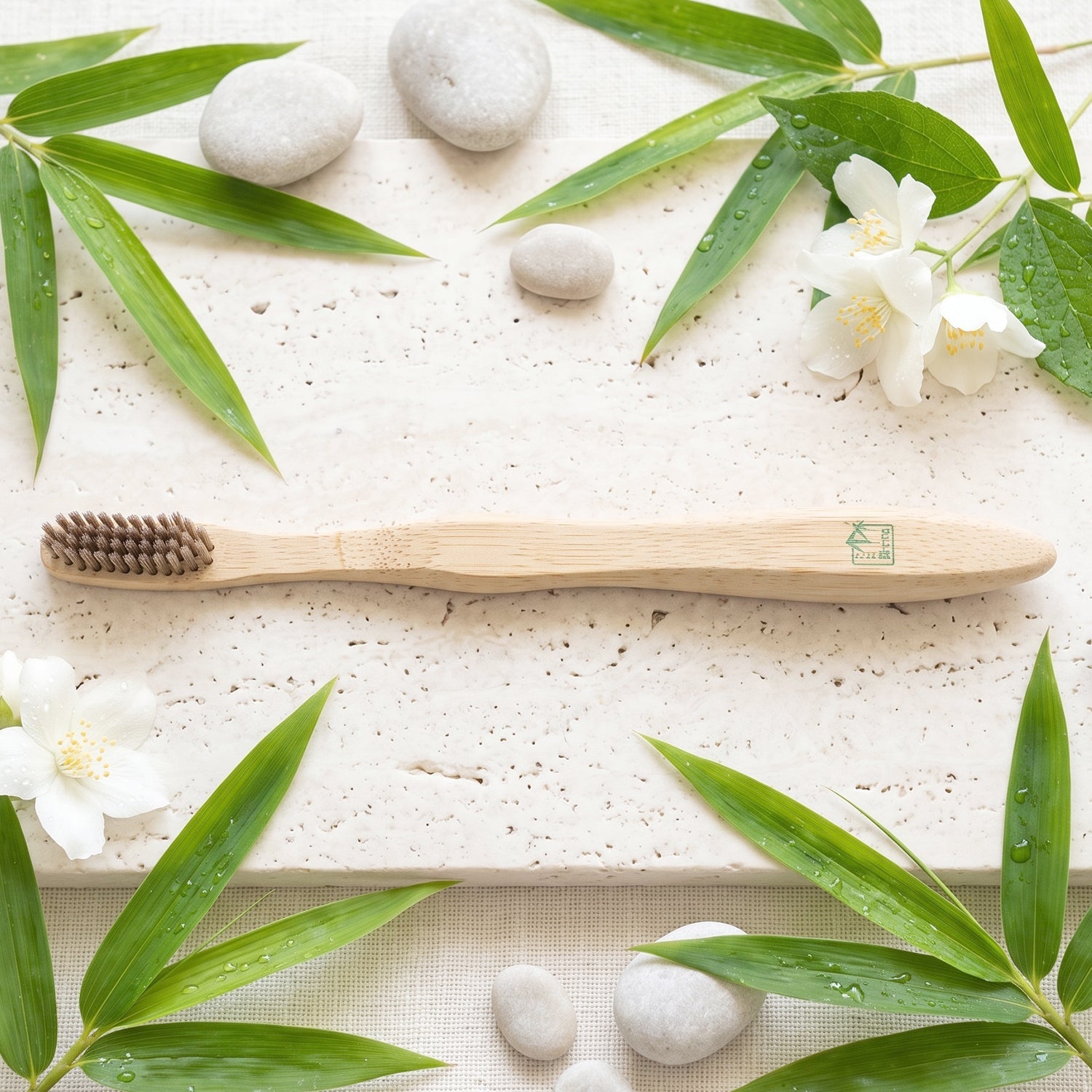 A bamboo toothbrush on a light stone surface, surrounded by green bamboo leaves, white flowers, and smooth grey stones.