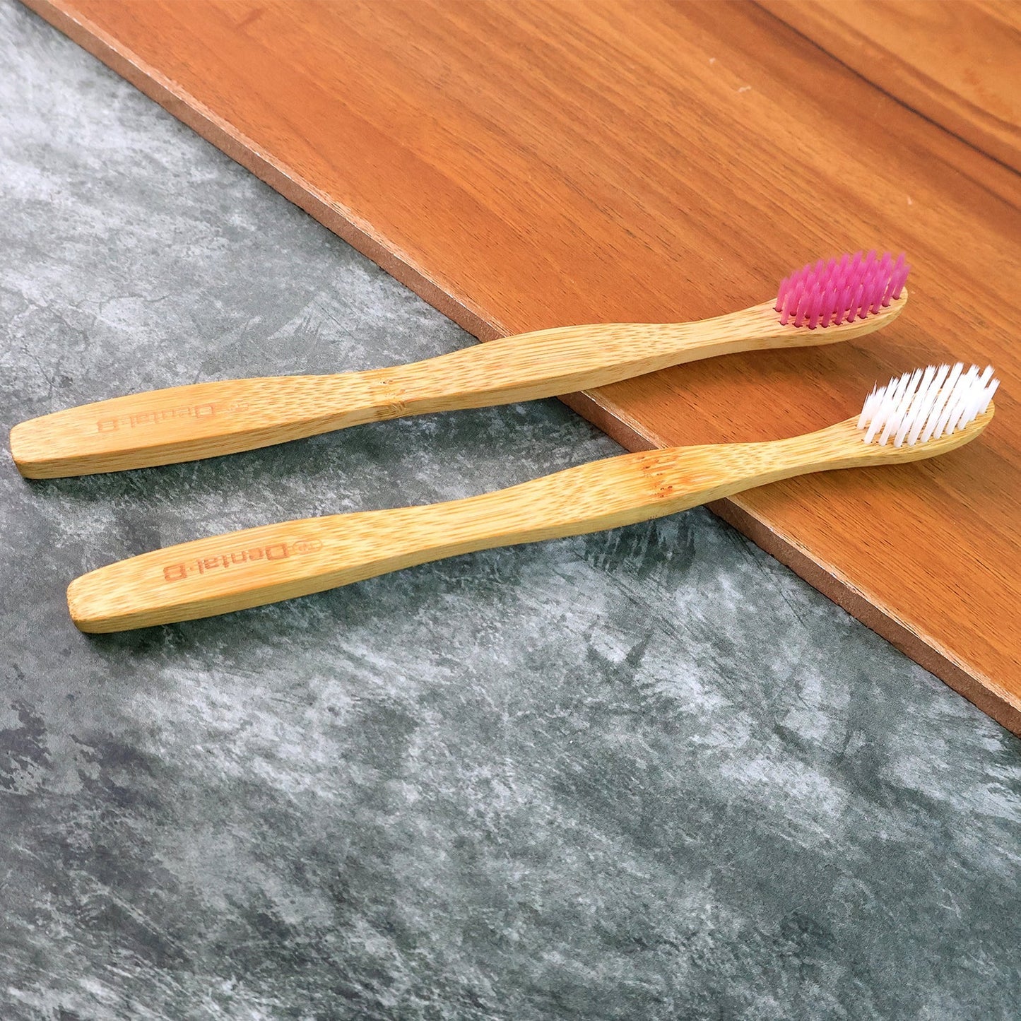Two bamboo toothbrushes, one with pink and one with white bristles, lying on a dark wooden and grey textured surface.
