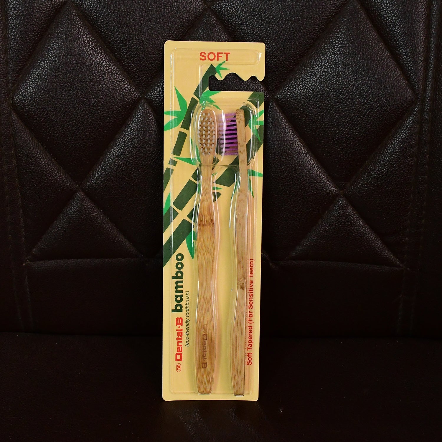 A two-pack of Dental B bamboo toothbrushes, one with brown and one with purple bristles, in yellow packaging against a dark