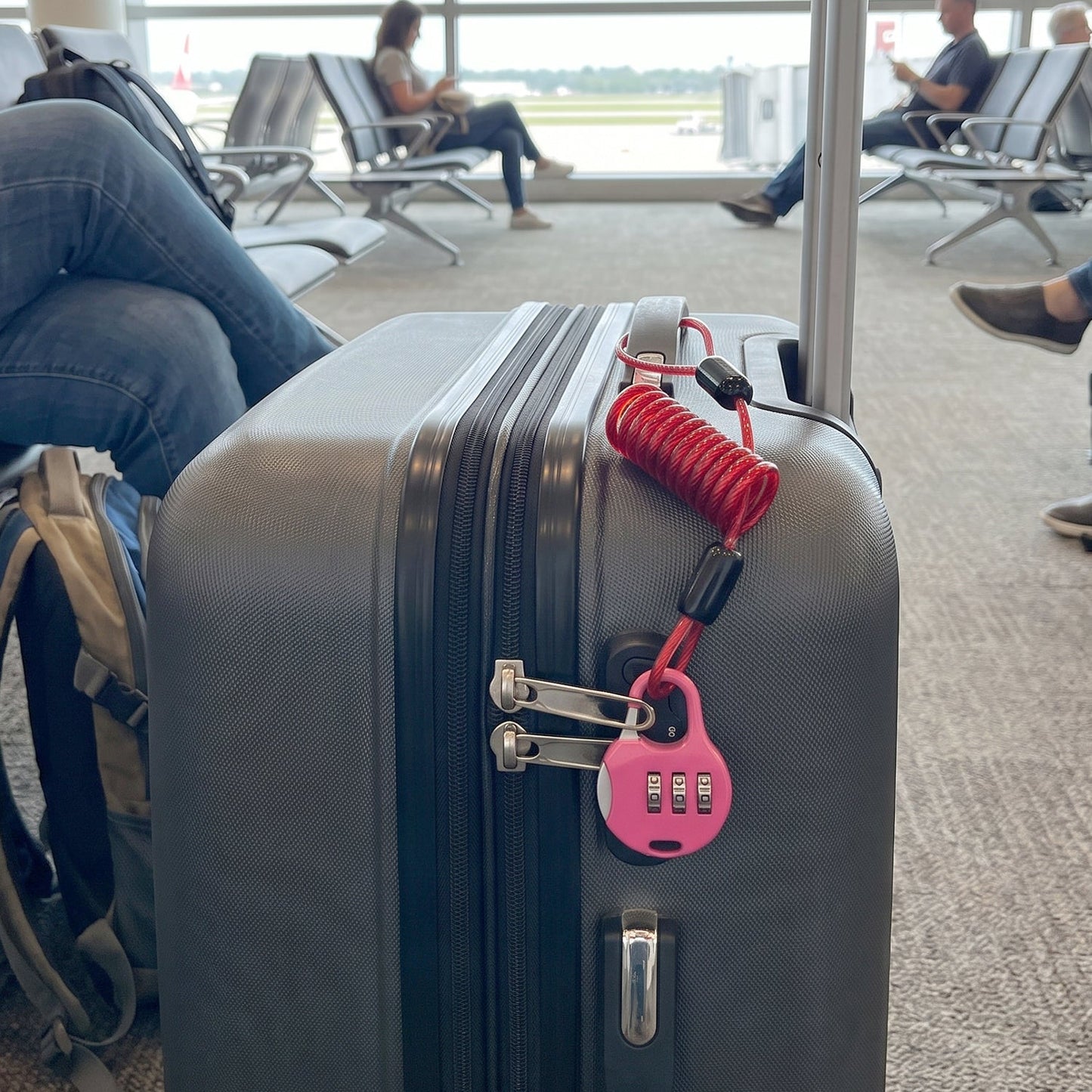 A pink combination lock with a red coiled cable secures a gray suitcase at an airport gate.