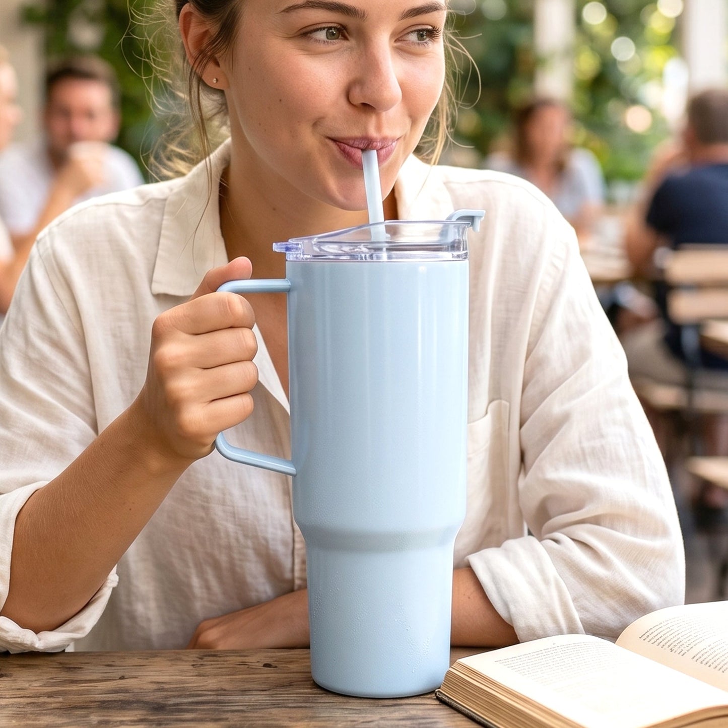 A woman sips from a light blue travel tumbler with a straw at a wooden table outdoors.