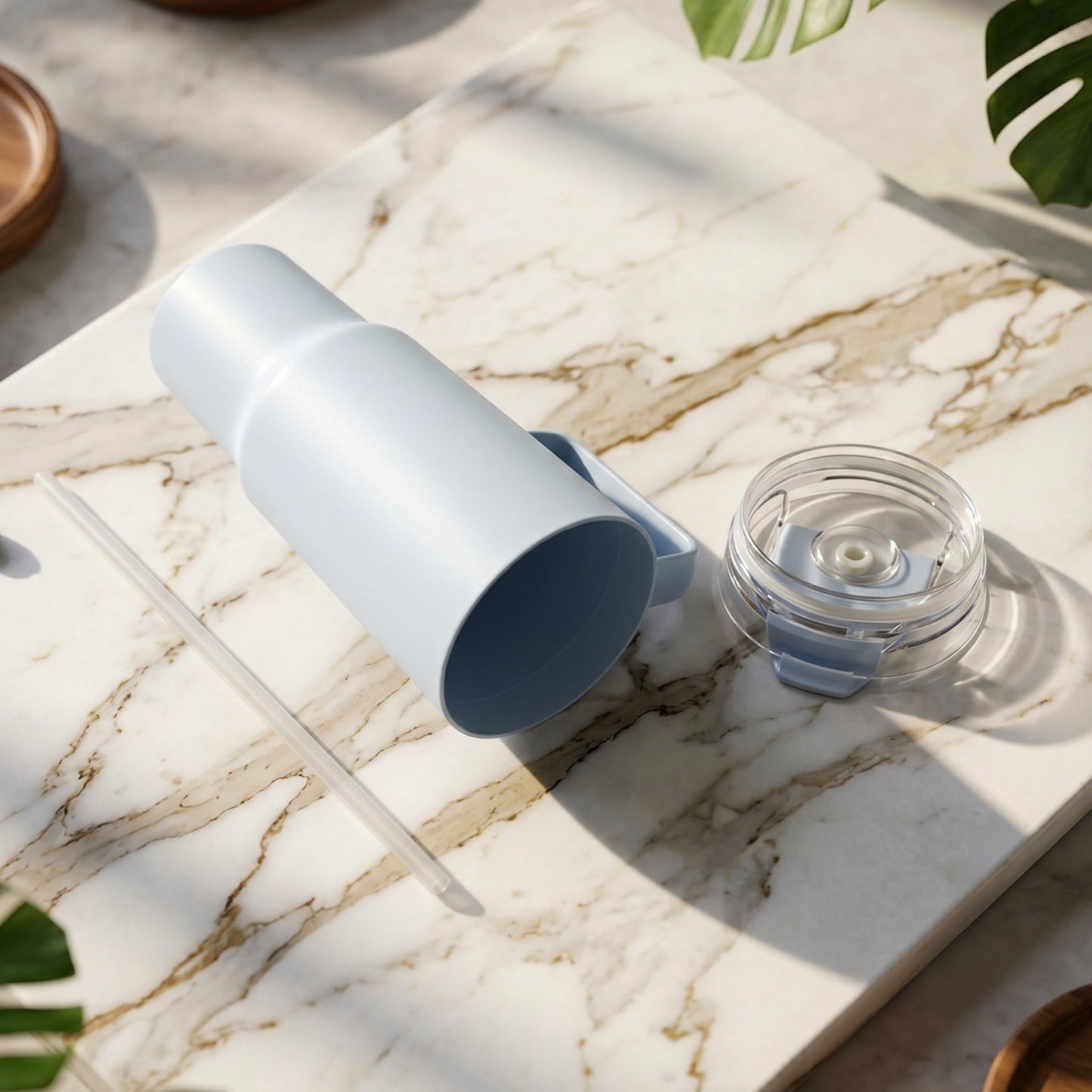A light blue travel tumbler, its clear lid, and a straw are laid on a white marble surface with green leaves.
