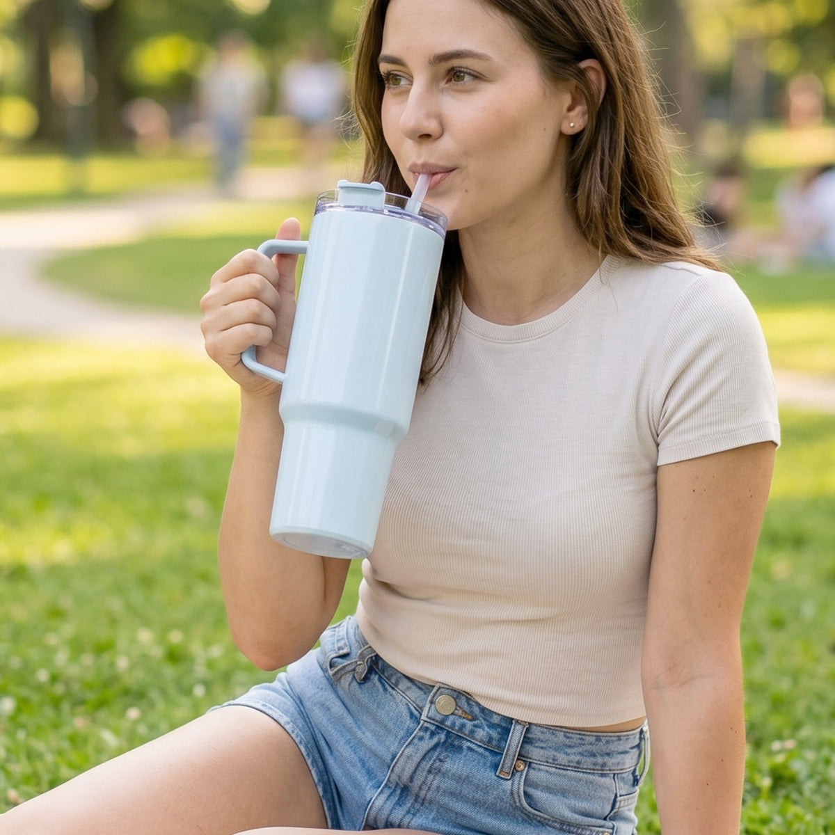 A woman in a t-shirt and shorts sips from a light blue travel tumbler while sitting on green grass.