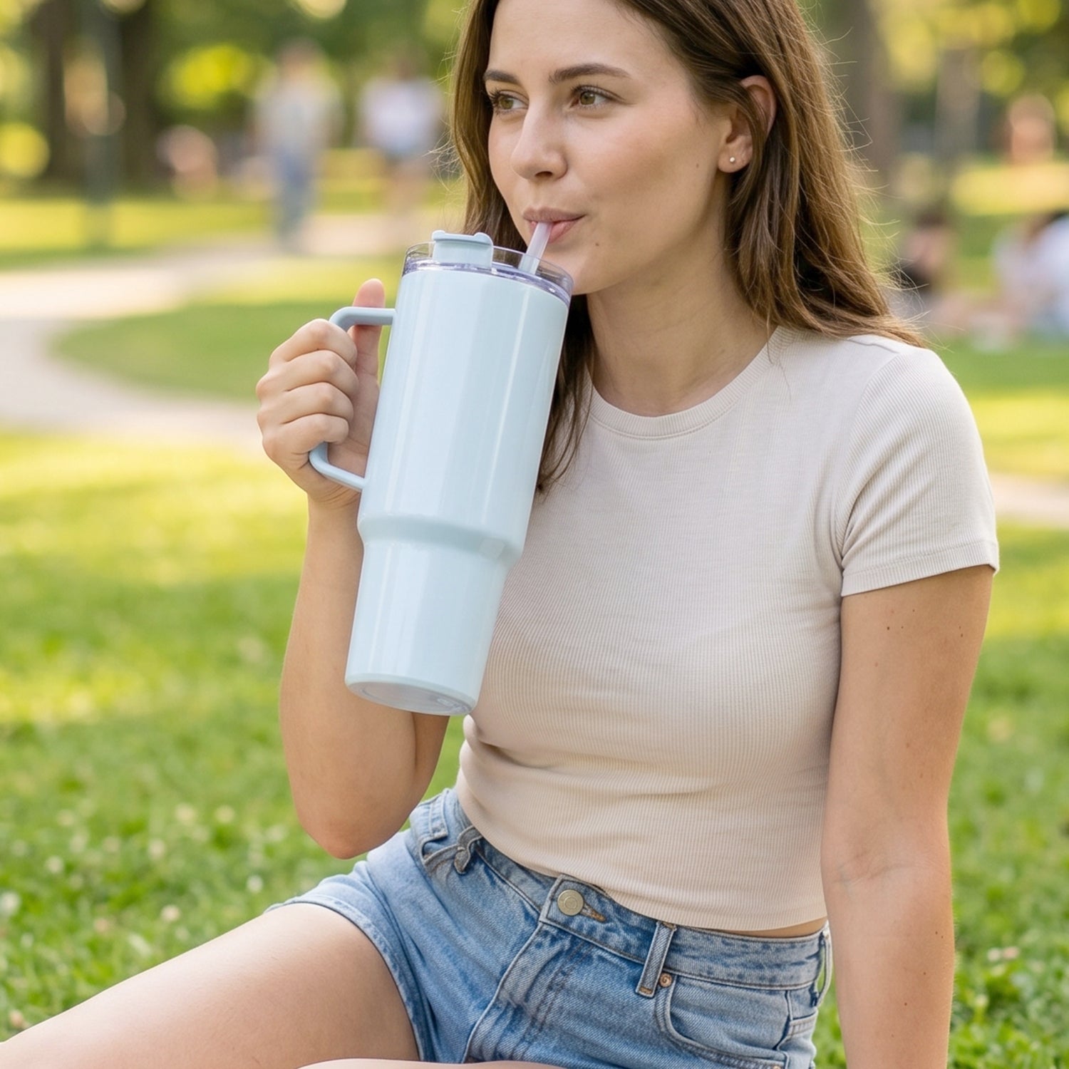 A woman in a t-shirt and shorts sips from a light blue travel tumbler while sitting on green grass.