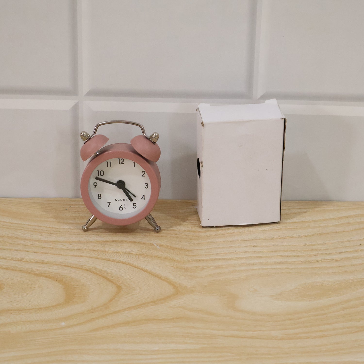 Pink twin bell alarm clock next to a small white open box on a light wooden surface with white tiled wall.